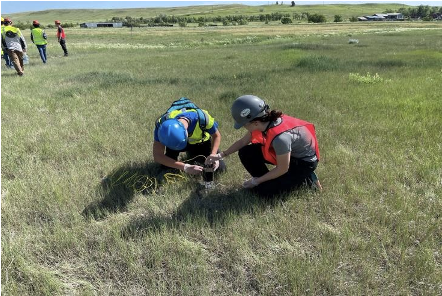 Two emergency responders wearing helmets and safety vests are kneeling on grass in a field, handling equipment connected to a monitor or device, with a few other people in the background and open landscape.