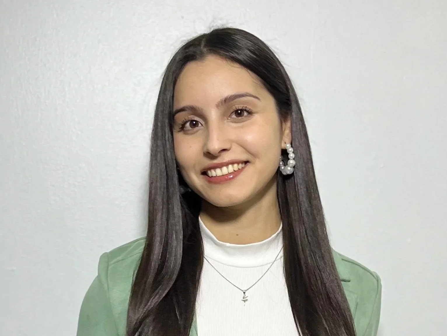 A young woman with long dark hair, smiling, wearing a white turtleneck, green blazer, large pearl earrings, silver necklace with a cross pendant, standing against a plain light-colored wall.