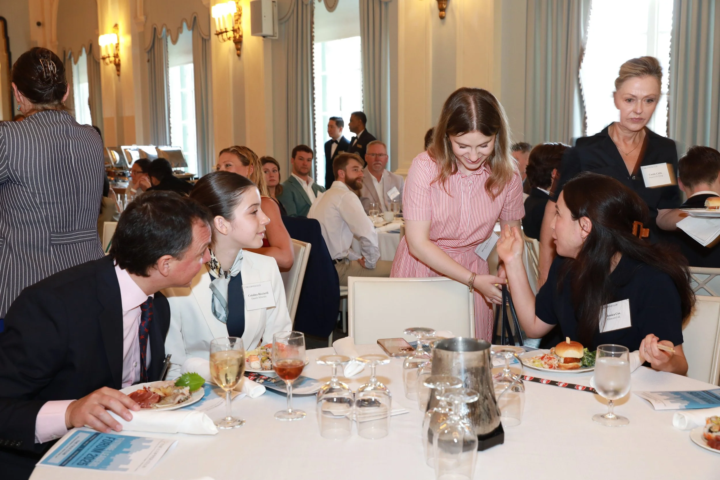 People attending a formal event or conference seated at dining tables, with women serving food and engaging in conversation, while some guests are standing in the background in a well-lit elegant room.