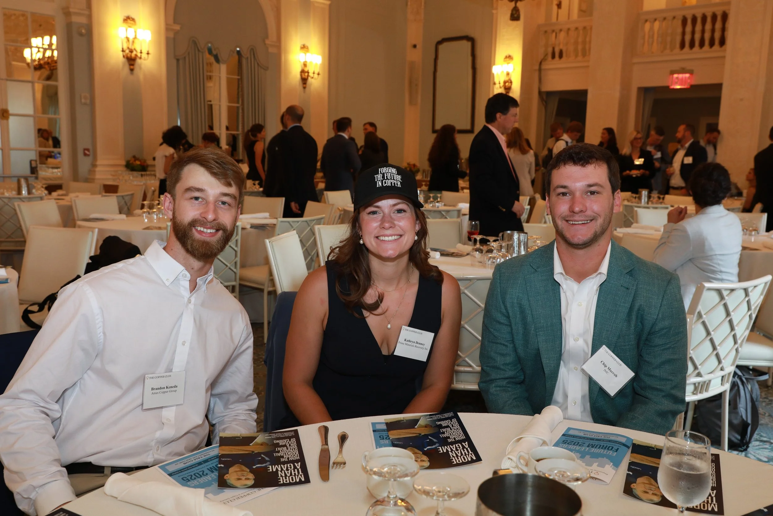 Three people smiling at a formal event, sitting at a table set with plates, silverware, and brochures. The woman in the middle is wearing a black dress, a black cap with white text, and earrings, while the men on either side are dressed in white shir