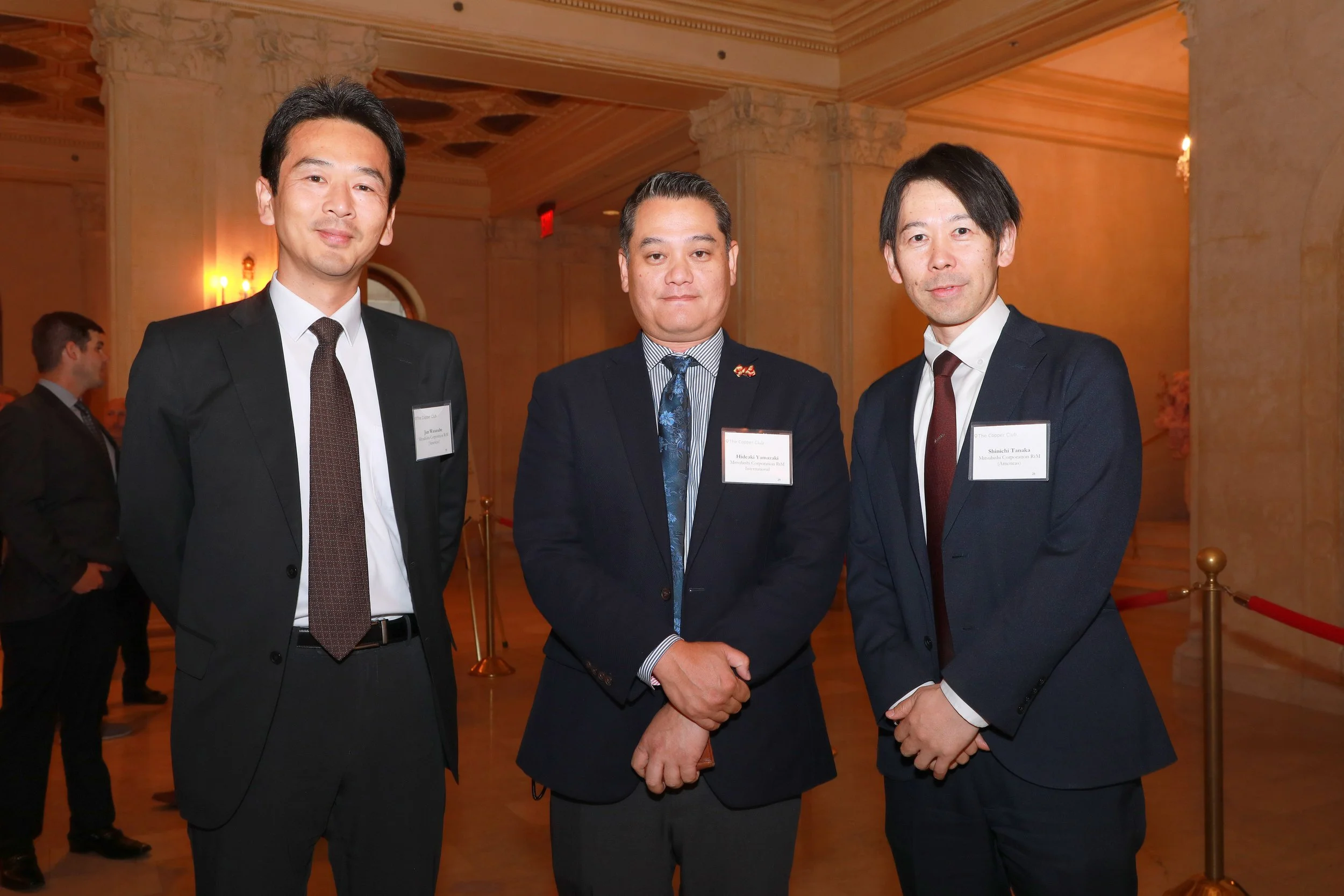 Three men in suits at a formal event, standing in a line, with one handshake happening between the man in the middle and the man on the right.