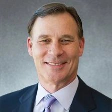 Professional headshot of a man in a suit smiling against a gray background.