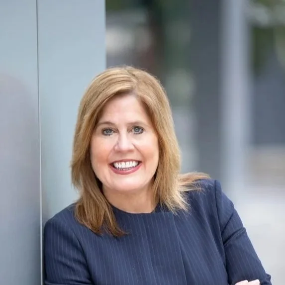 A woman with shoulder-length blonde hair, smiling, wearing a navy blue pinstripe blazer, standing outdoors next to a glass building wall.