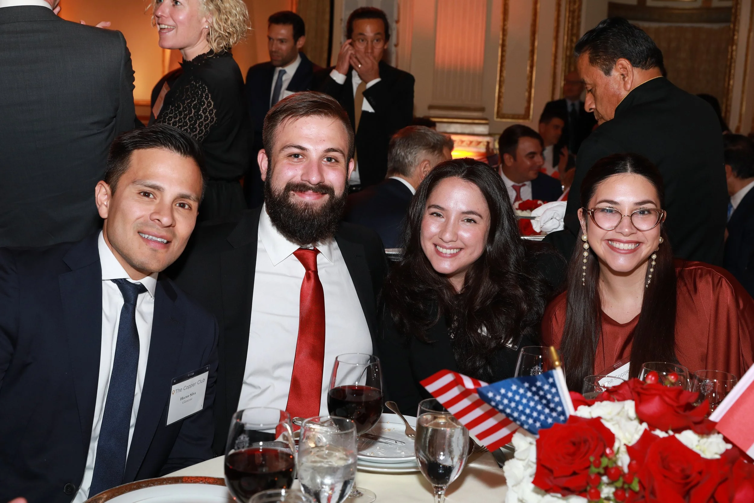 Group of people smiling and sitting at a formal dinner table decorated with red and white flowers, American flags, and wine glasses, at an elegant event or banquet.