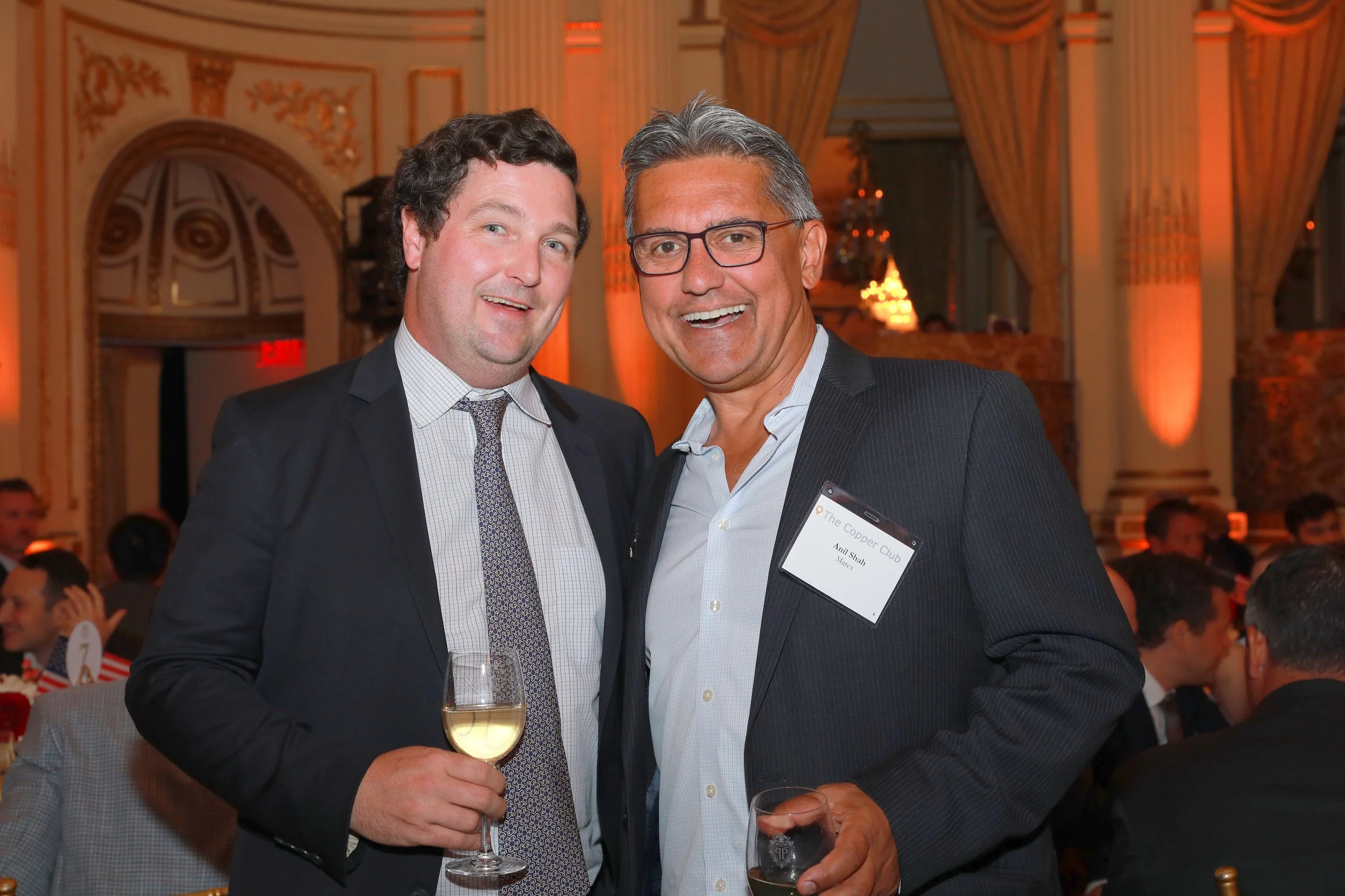 Two men dressed in suits at a formal event, smiling and holding drinks, in a grand decorated venue with ornate walls and drapes.