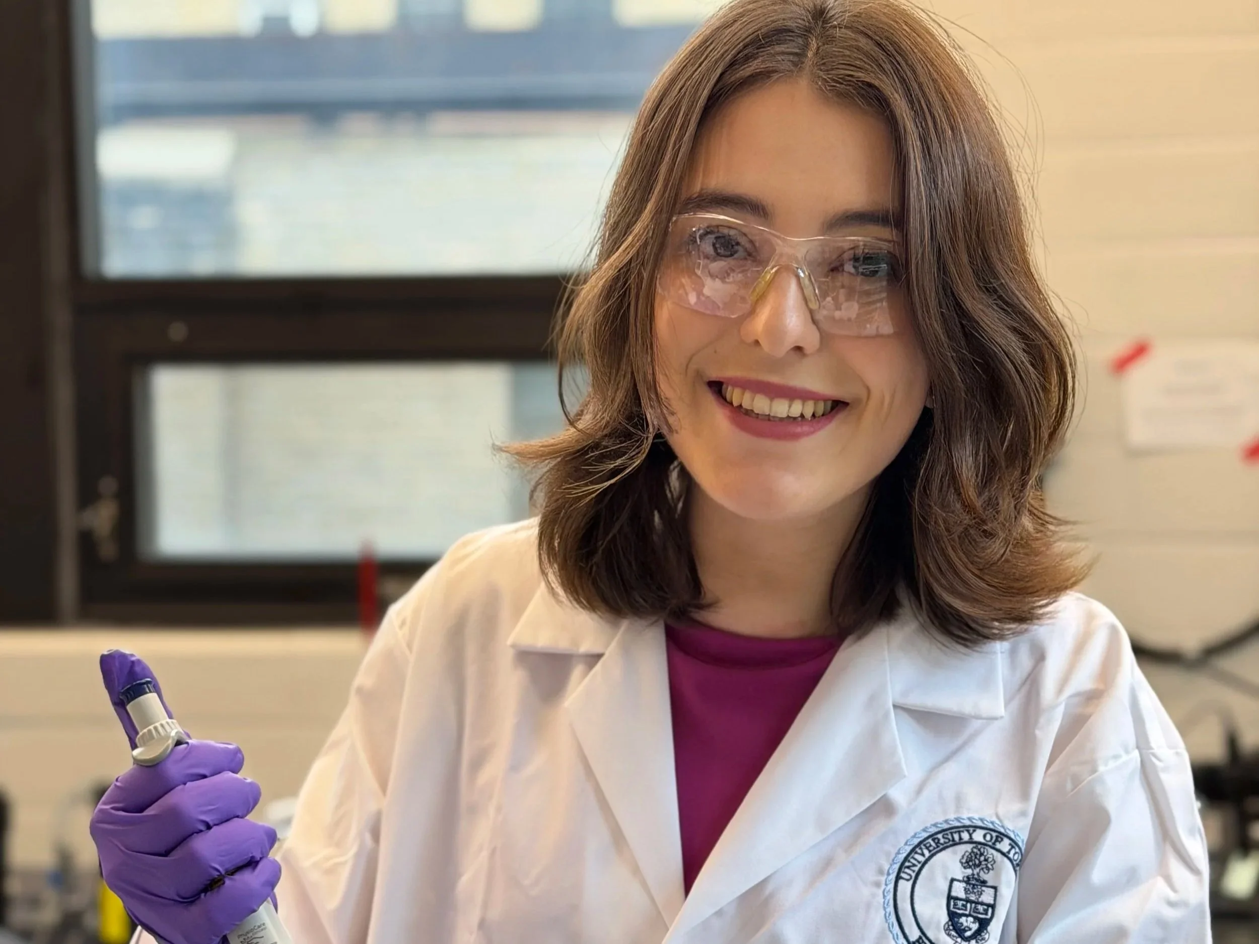 A woman with brown hair and wearing safety glasses and a white lab coat with a university logo, smiling and holding a purple marker, working in a laboratory.