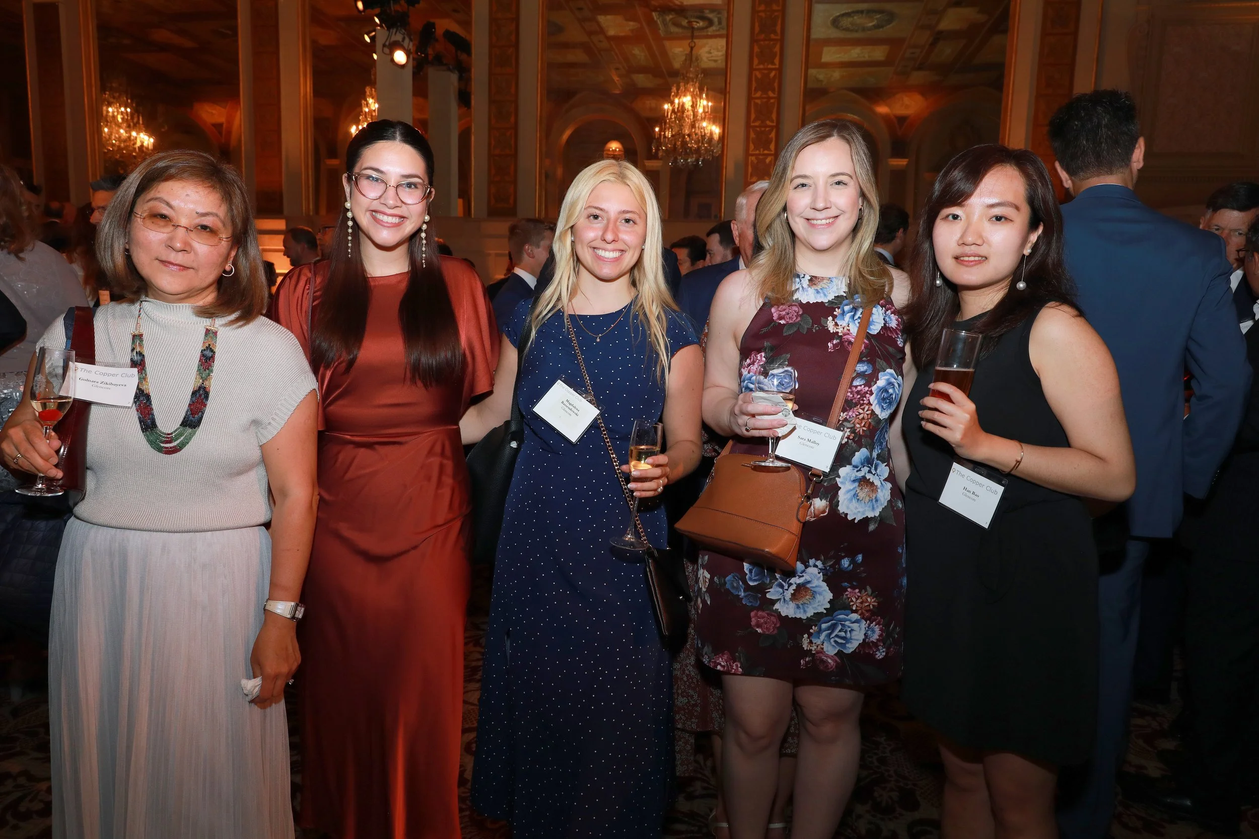 Group of five women smiling at a formal event, holding glasses of champagne, in an ornate banquet hall with chandeliers.