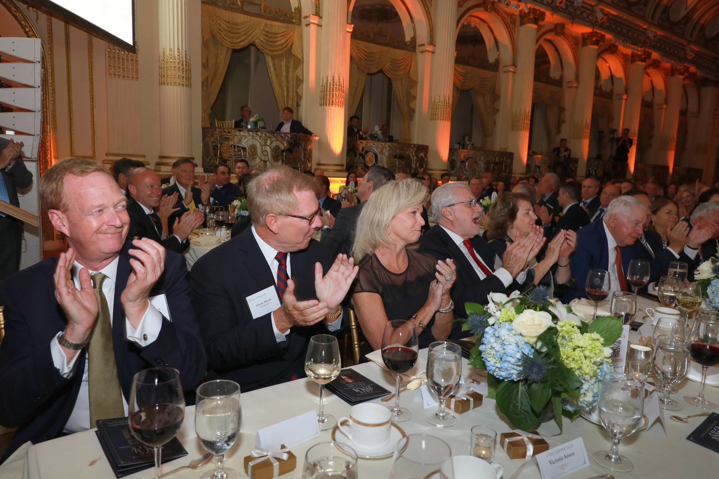 A formal banquet with a large group of people clapping and smiling at a table with floral centerpieces and wine glasses in a grand, ornately decorated hall with tall columns and gold accents.