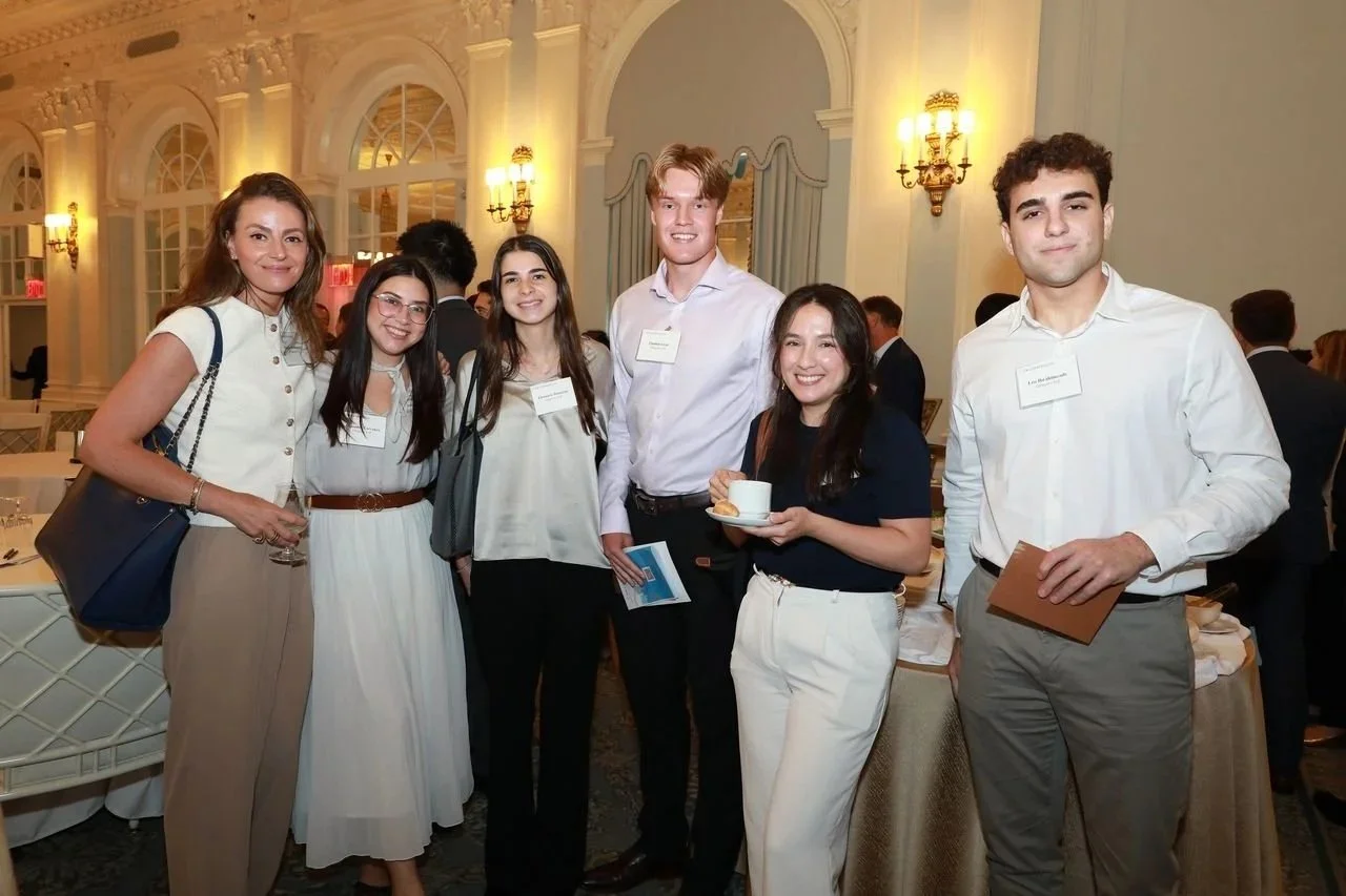 Group of six young adults at a formal event, smiling, in an elegant room with ornate decor and warm lighting.