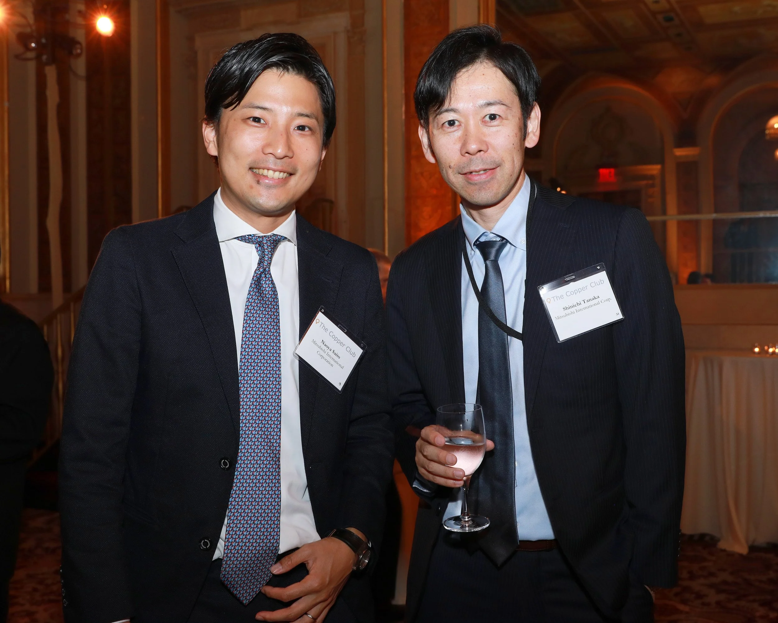 Two men in formal attire at a social event, one smiling broadly and the other holding a glass of water, standing in a warmly lit elegant indoor setting with ornate decor.