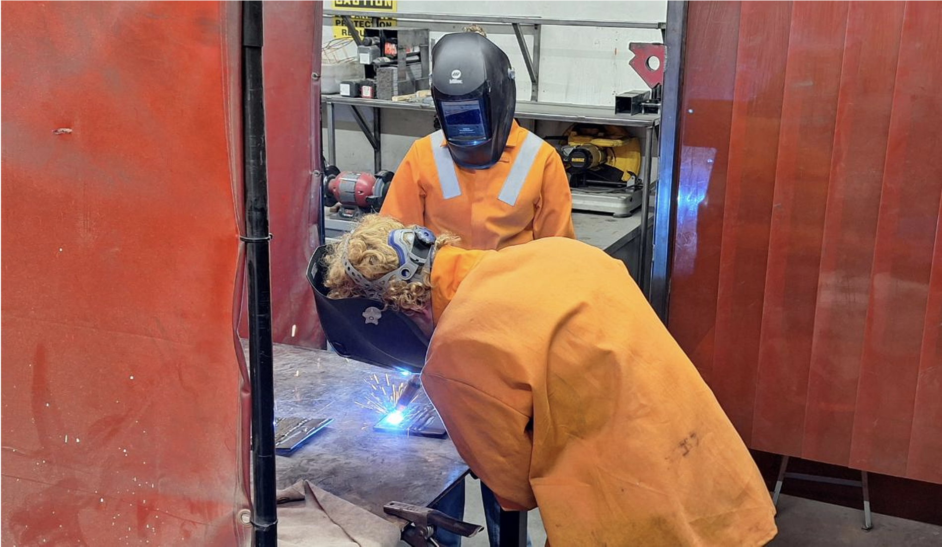 Two workers in orange protective clothing welding metal inside a workshop surrounded by tools and equipment. One worker is bending over, actively welding, while the other stands nearby, watching.