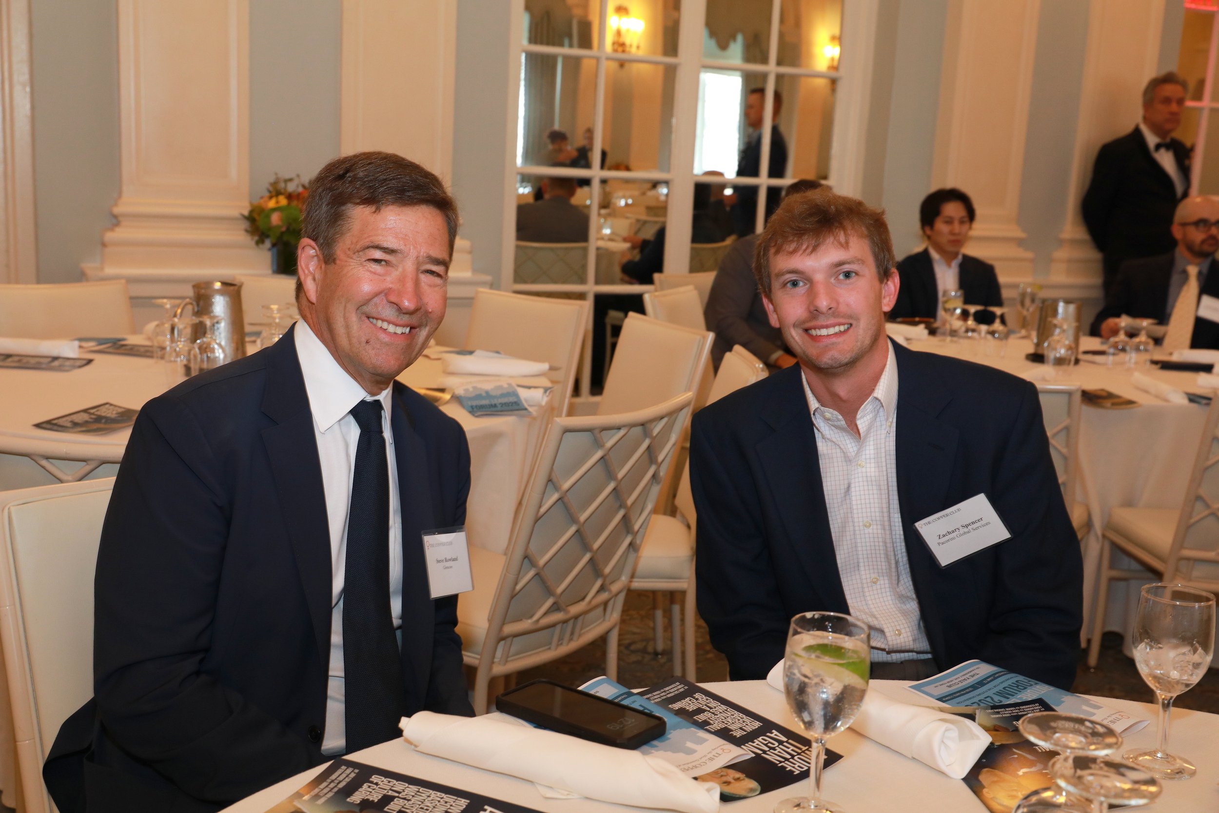Two men in formal attire sitting at a banquet table during a professional event. The man on the left has brown hair, a suit, and a name tag, smiling at the camera. The man on the right has light brown hair, a checked shirt, and a suit jacket, also sm