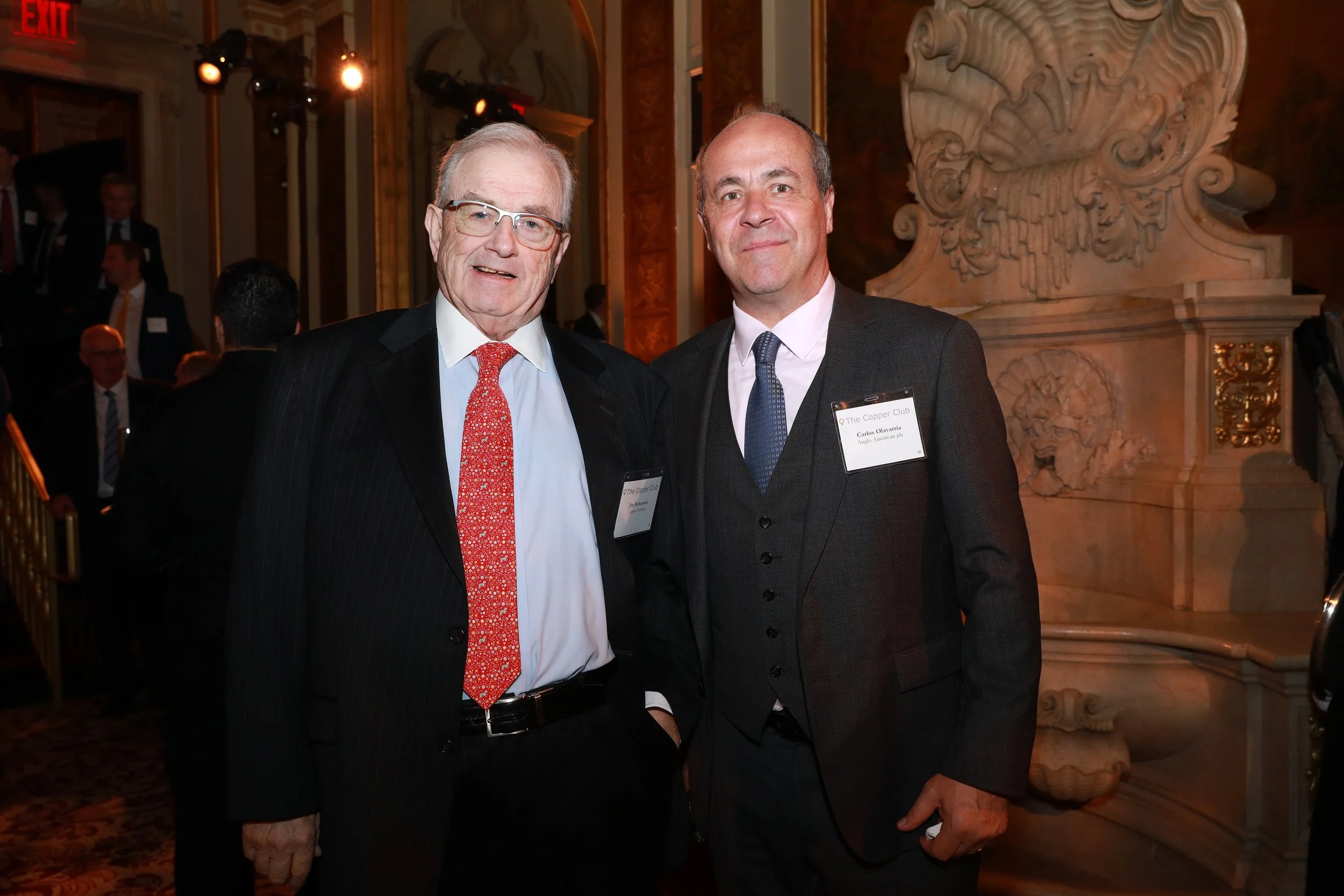 Two men in suits attending an event, standing in a decorated venue with ornate wall art and lighting, smiling at the camera.