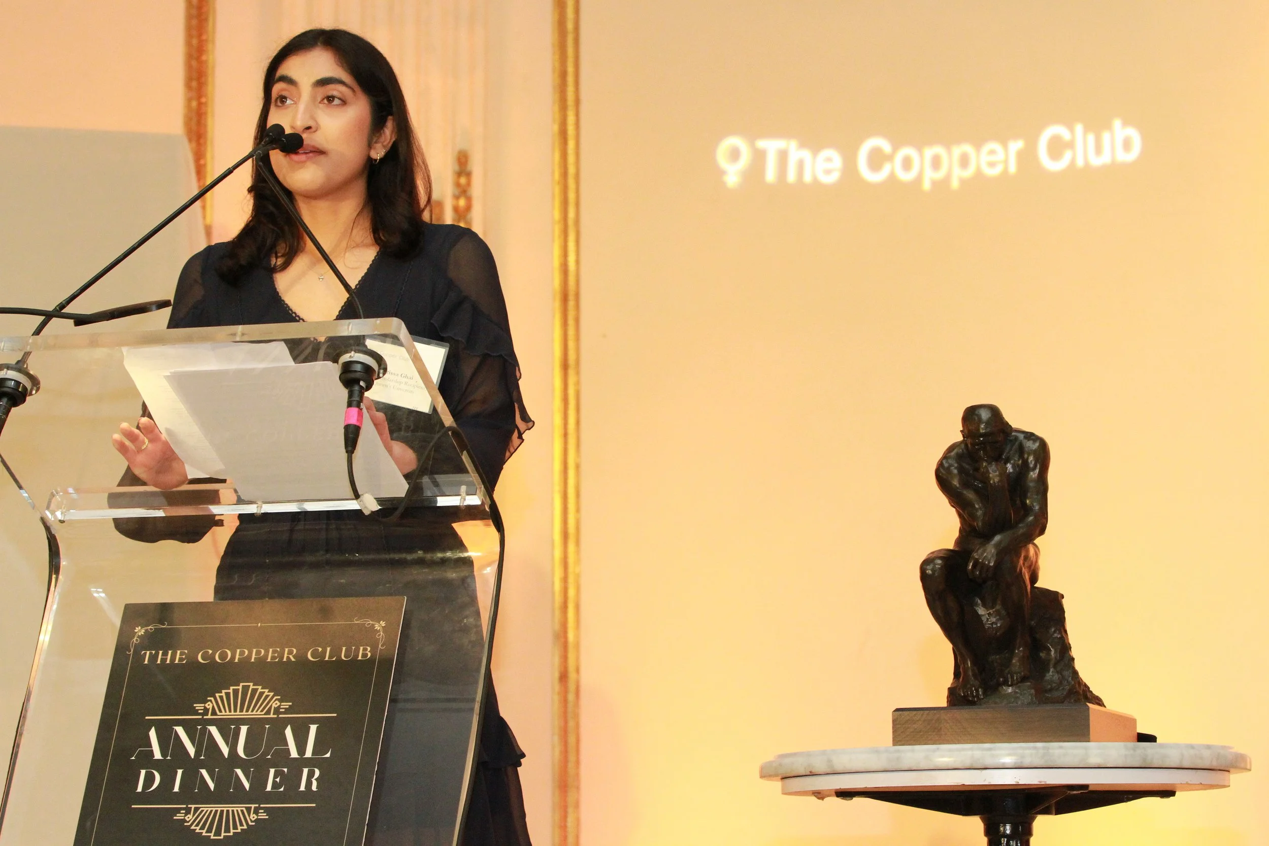 A woman speaking at a podium during an event with a sign that reads 'The Copper Club' and 'Annual Dinner'. There is a sculpture of a person sitting with their head bowed on a table beside her.
