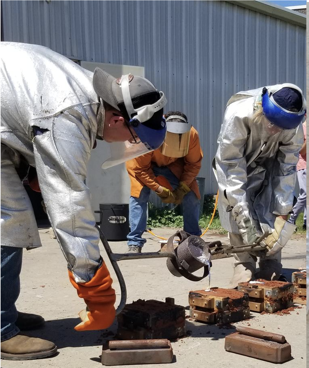 Workers in protective gear, including helmets, face shields, jackets, and gloves, are conducting a metal casting or melting operation outdoors. They are handling molten metal poured into molds.