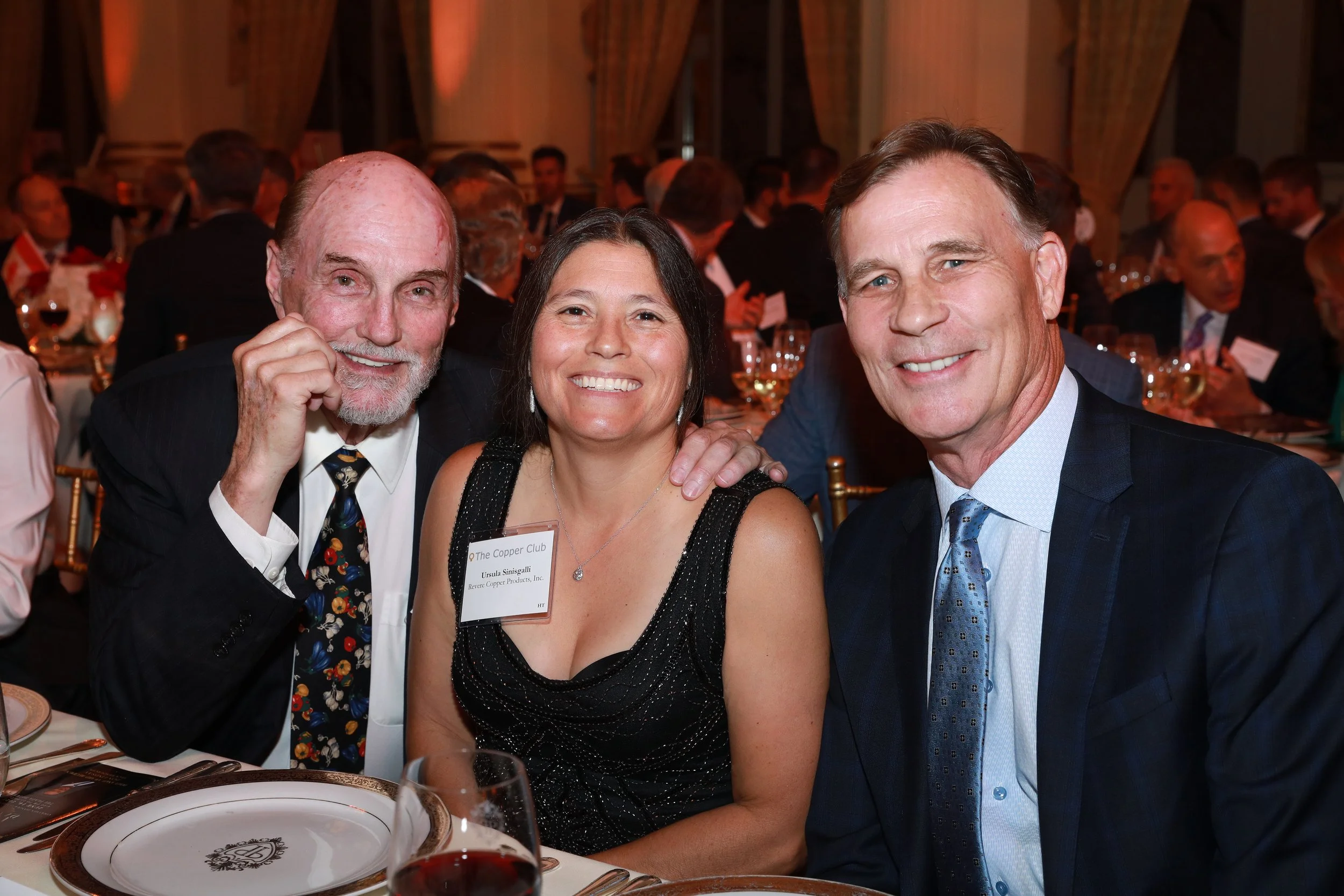 Three people smiling at a formal dinner event, sitting at a table with wine glasses and tableware.