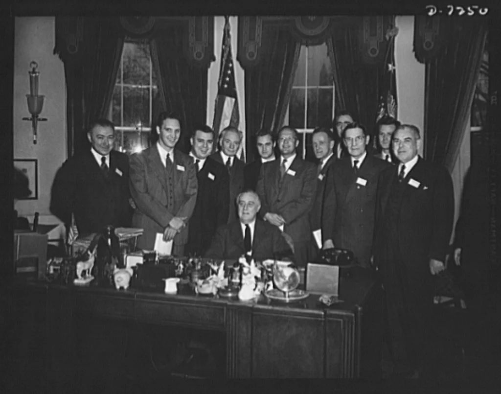 Black and white photo of a group of men in suits and ties gathered around a desk, with one man seated in the center. The setting appears to be an office or formal room, with curtains, a window, and an American flag in the background.