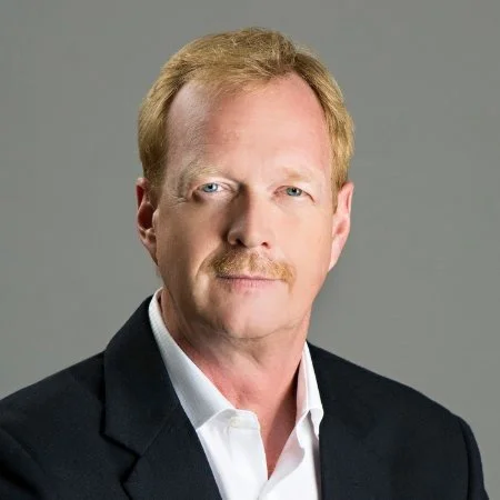 A middle-aged man with red hair, a mustache, and light-colored eyes, wearing a black blazer and white shirt, posing against a gray background.