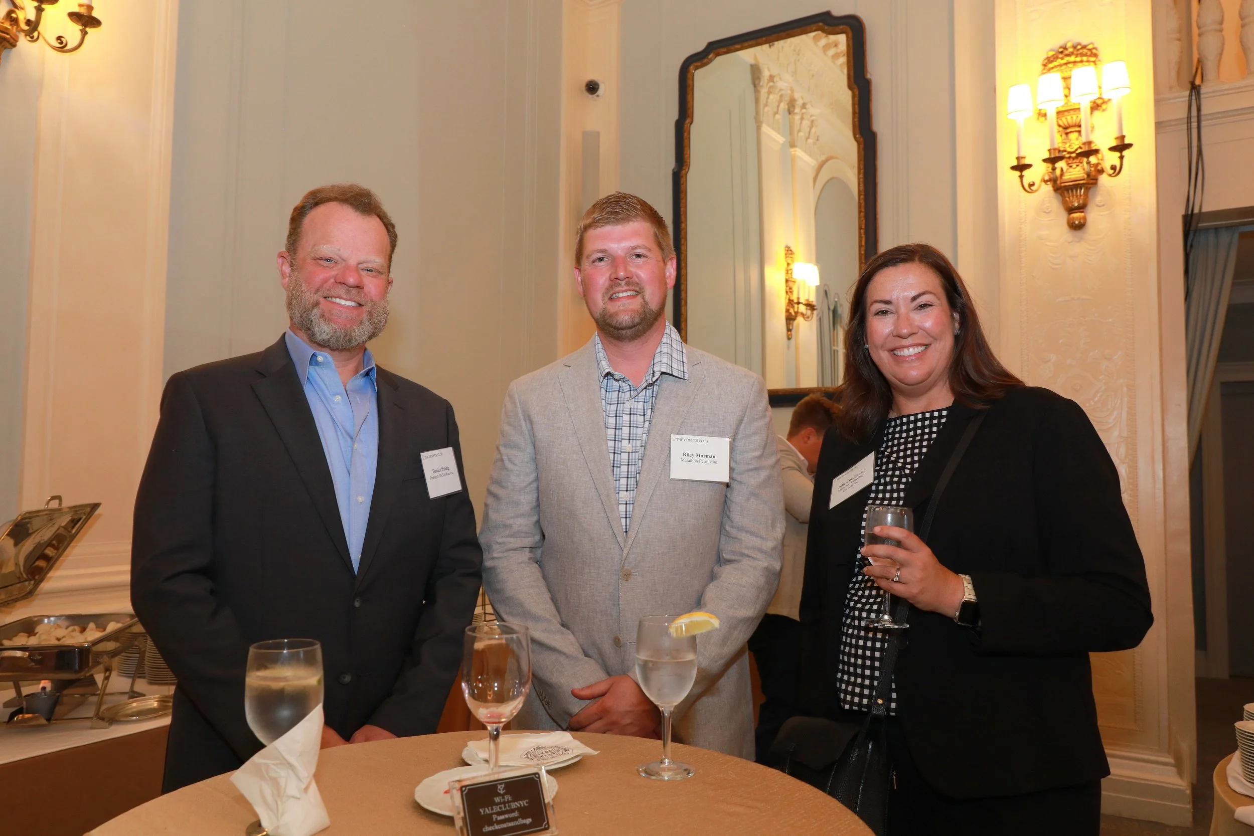Three people, two men and one woman, standing together at a formal event in a decorated room with large mirror and wall sconce lights. The man on the left has a beard and is wearing a black suit with a blue shirt. The man in the middle has short hair