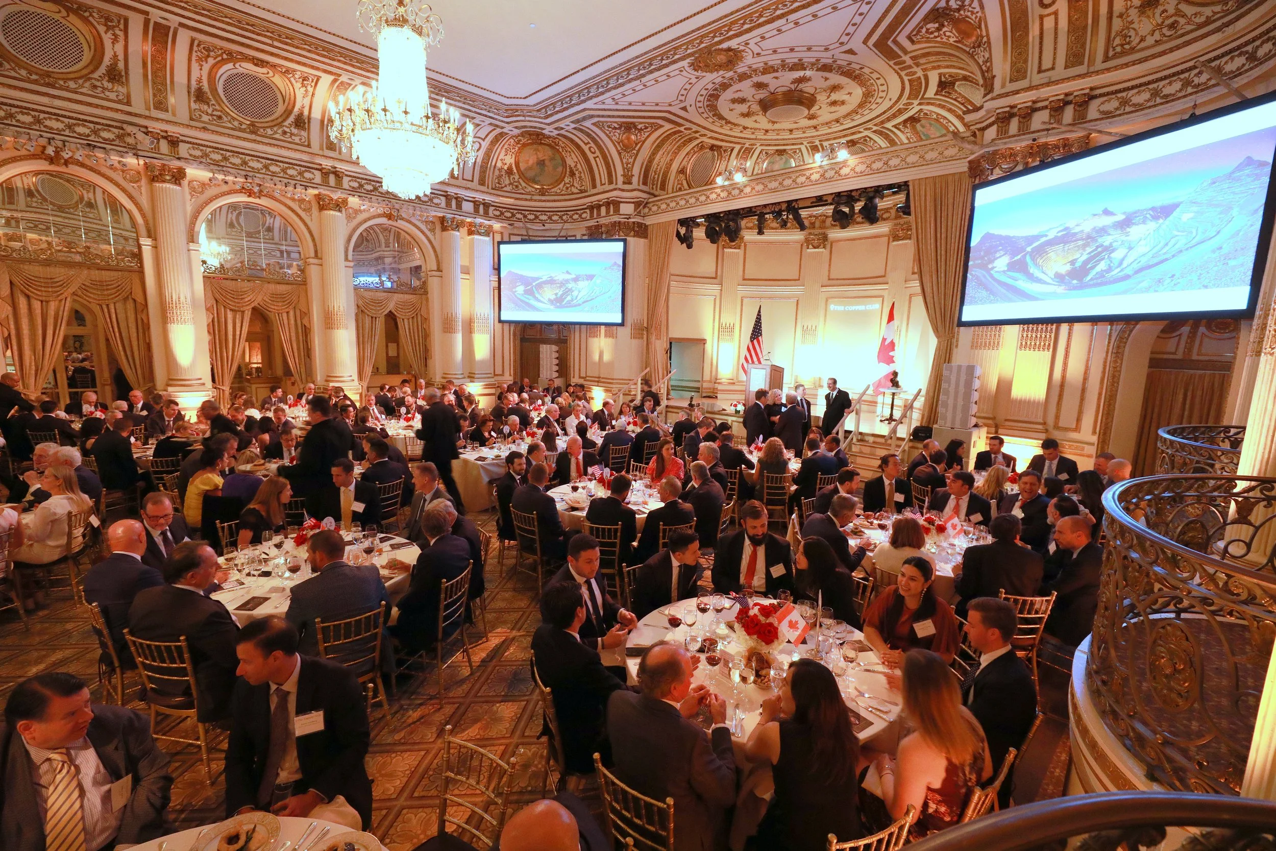 A large formal banquet hall filled with round tables of people dressed in business attire, engaged in conversation during an event. The room features ornate gold detailing, large chandeliers, and tall windows with gold curtains. Two large screens dis