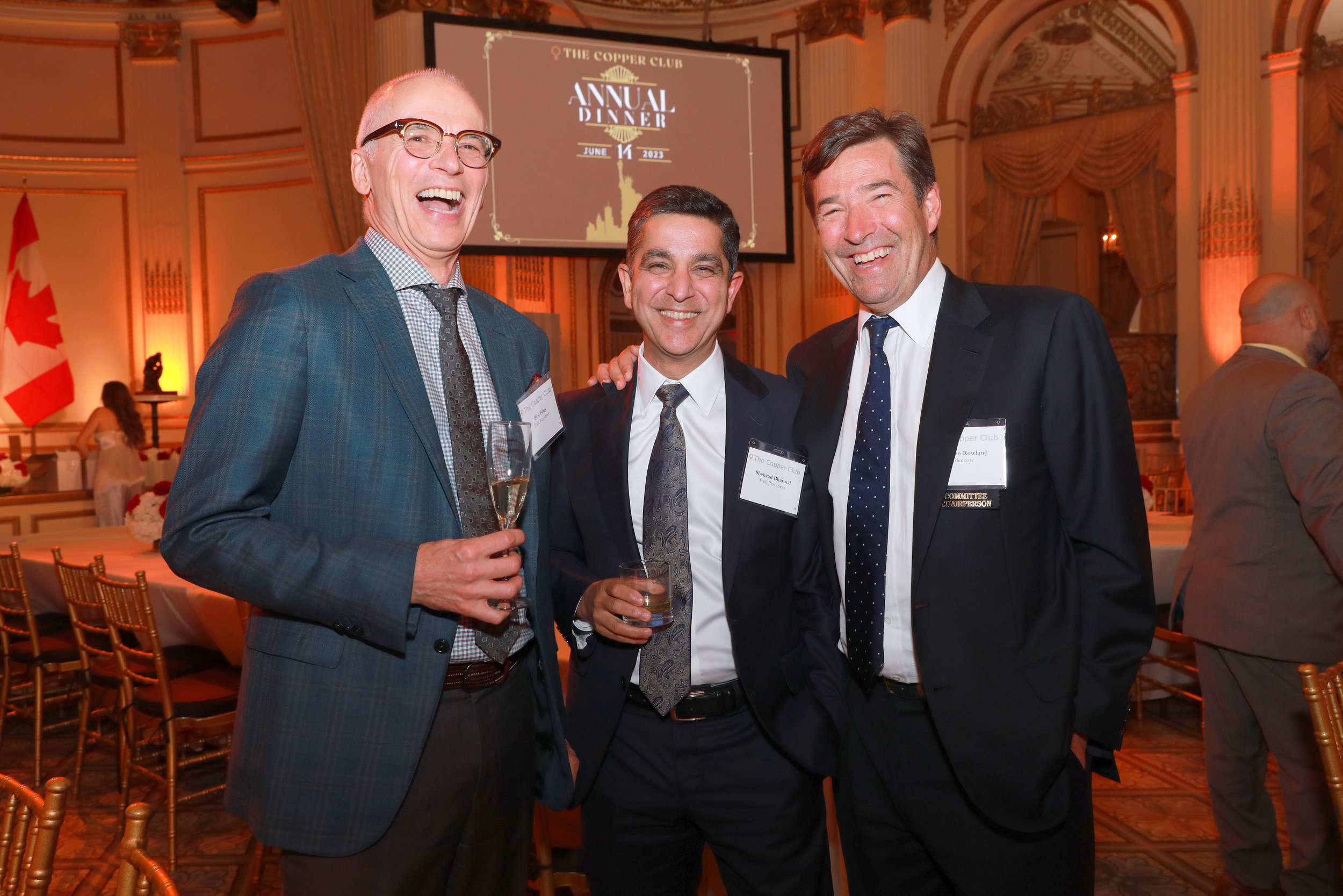 Three men in suits smiling and holding glasses at an indoor event with a large screen in the background displaying "The Copper Club, Annual Dinner, June 14, 2023" and a Canadian flag.