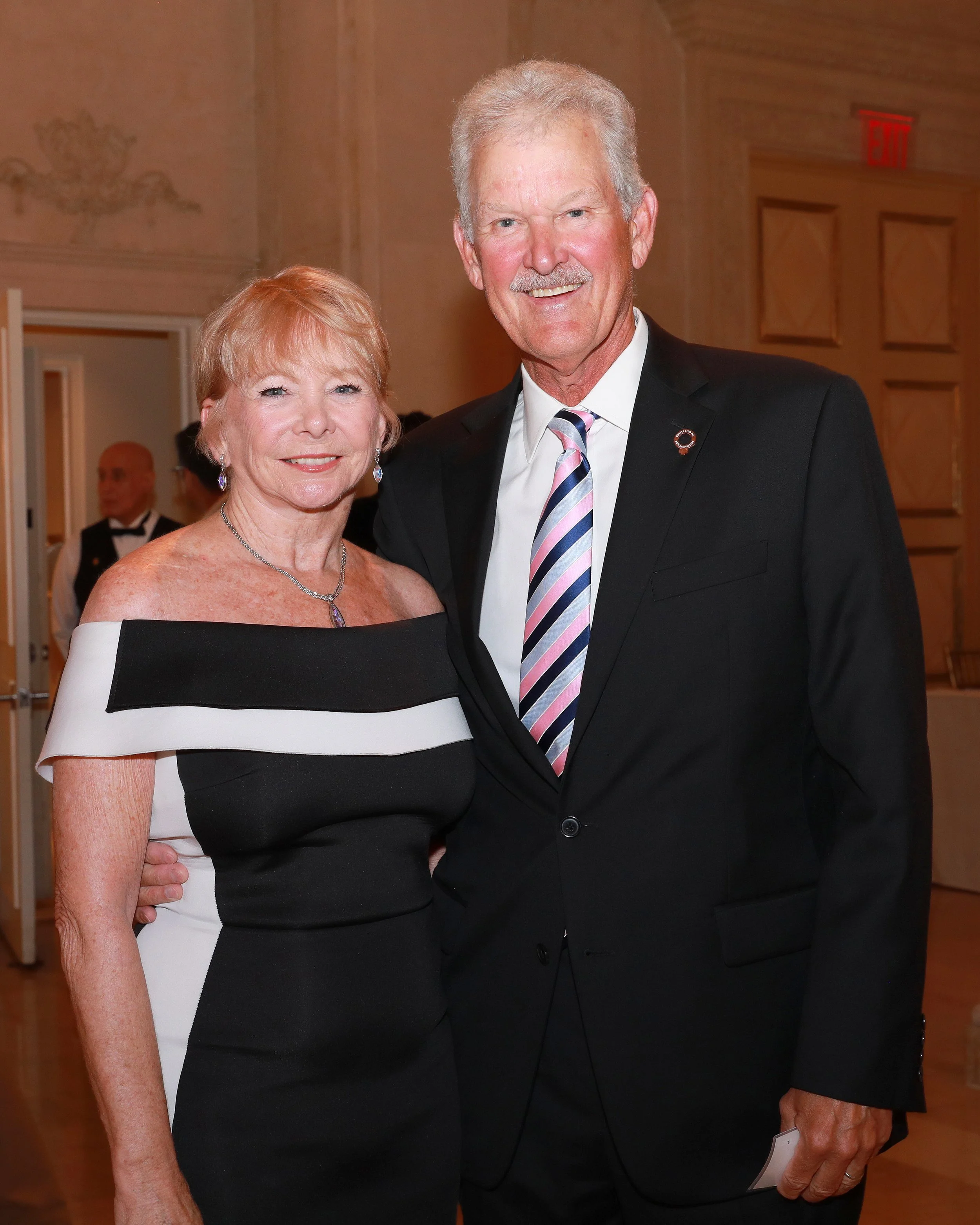 A man and a woman dressed formally, standing close together and smiling at a ballroom or event hall.