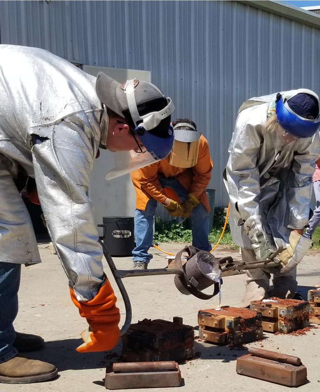 Workers in protective gear pouring molten metal into molds outdoors.