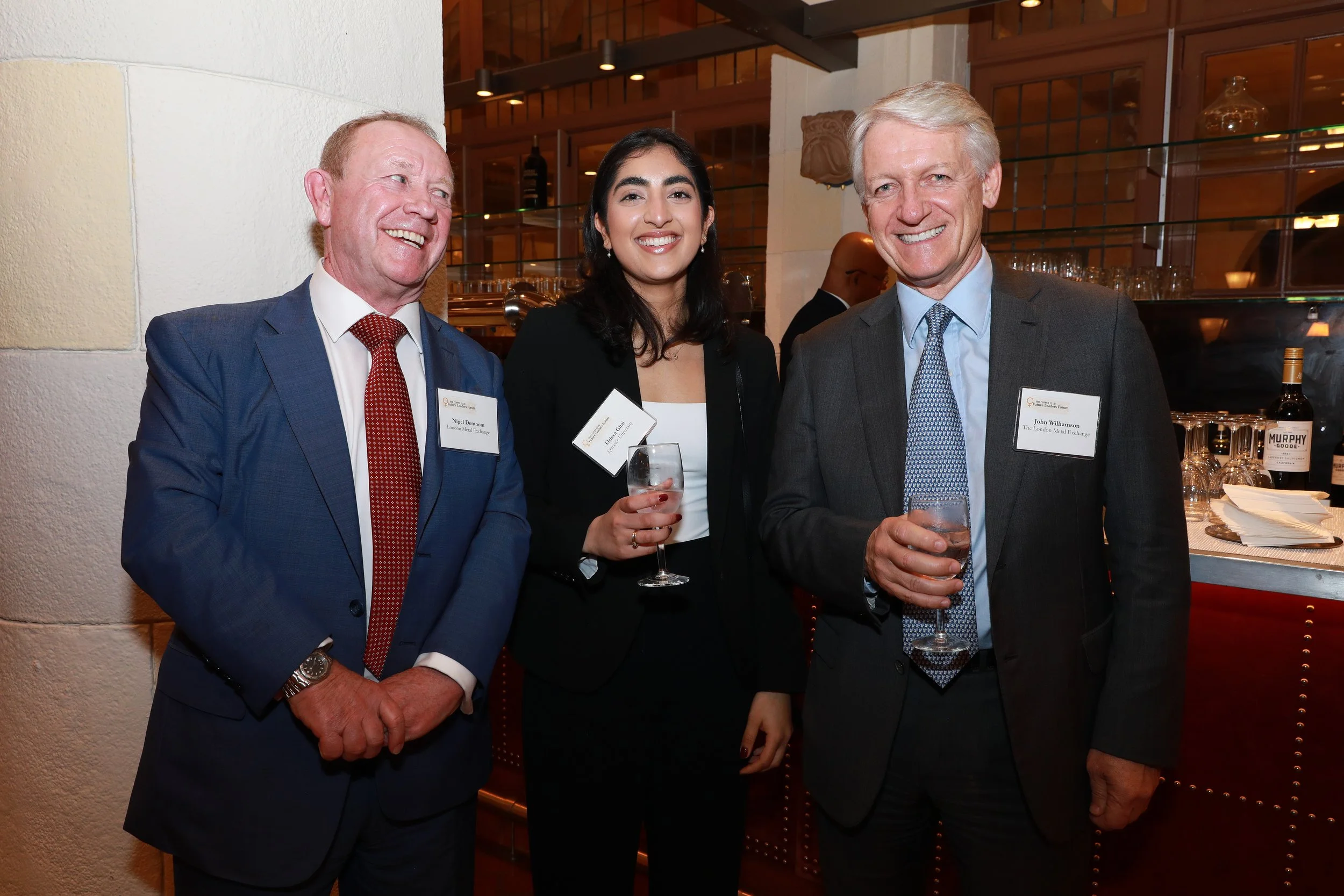 Three professionally dressed individuals at a social event, smiling and holding drinks, in an indoor setting with warm lighting and bar area in the background.