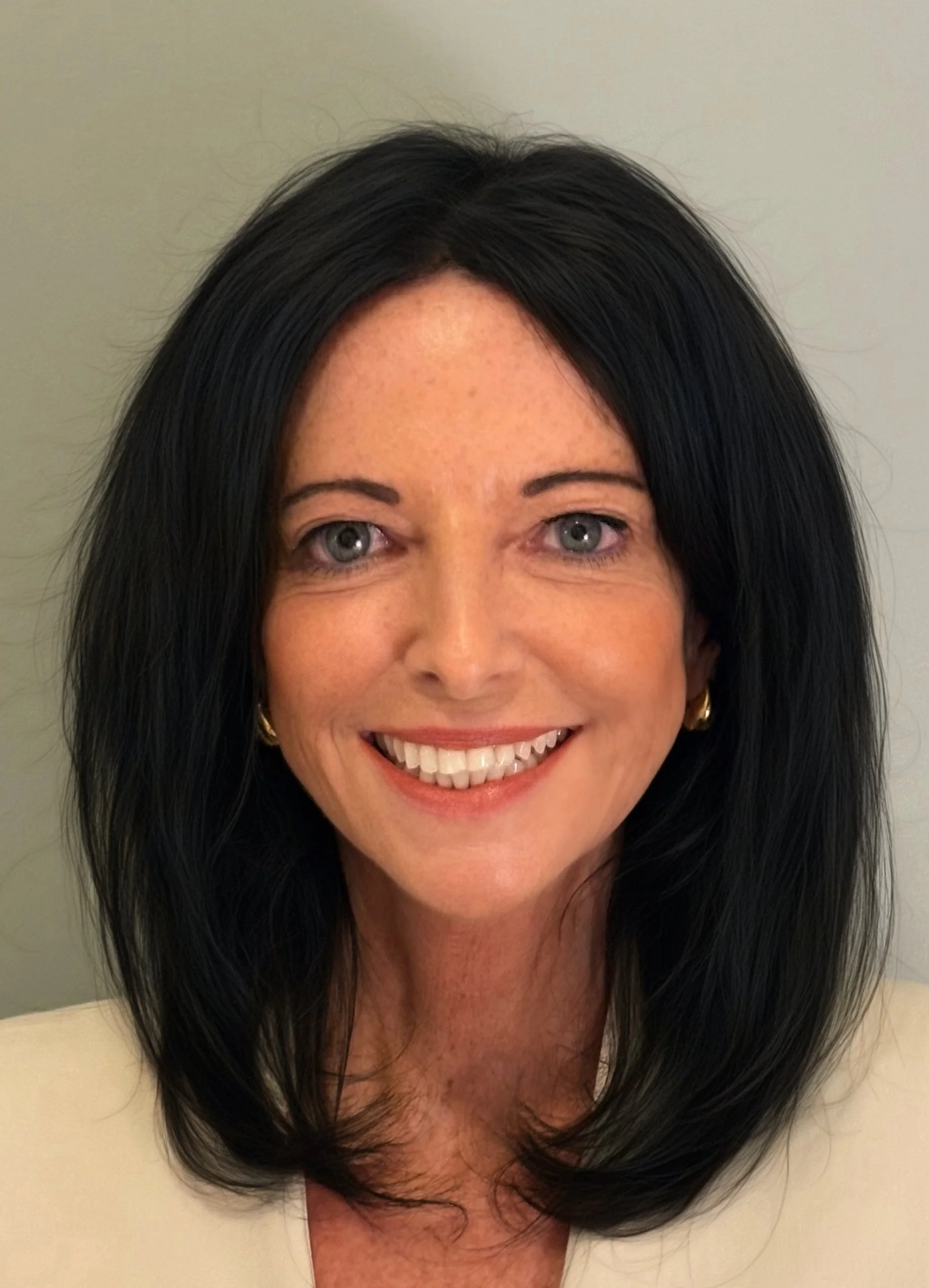 Close-up of a woman with long dark hair, wearing red lipstick, smiling, with a blurred outdoor background.