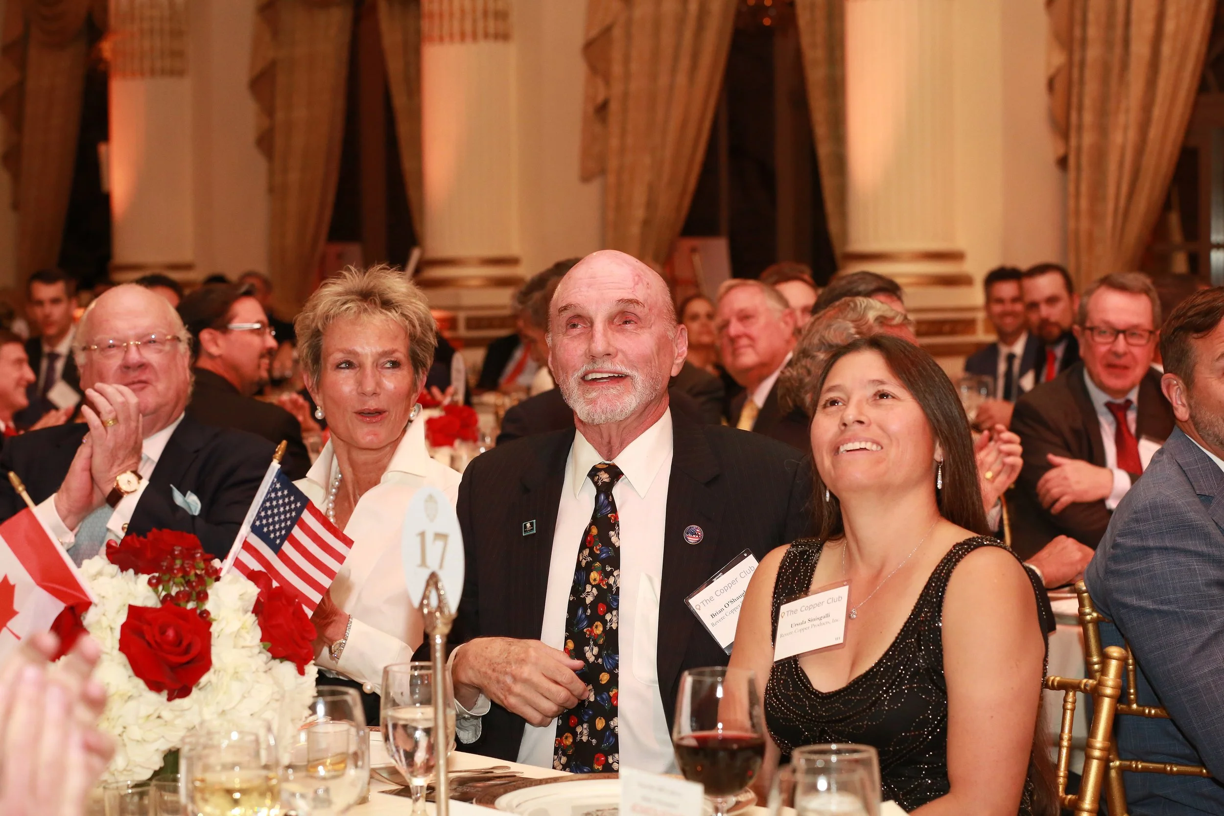 A group of people, dressed formally, seated at a banquet table with red, white, and blue decorations, including American flags and flowers, in an ornate room with tall columns and heavy curtains.