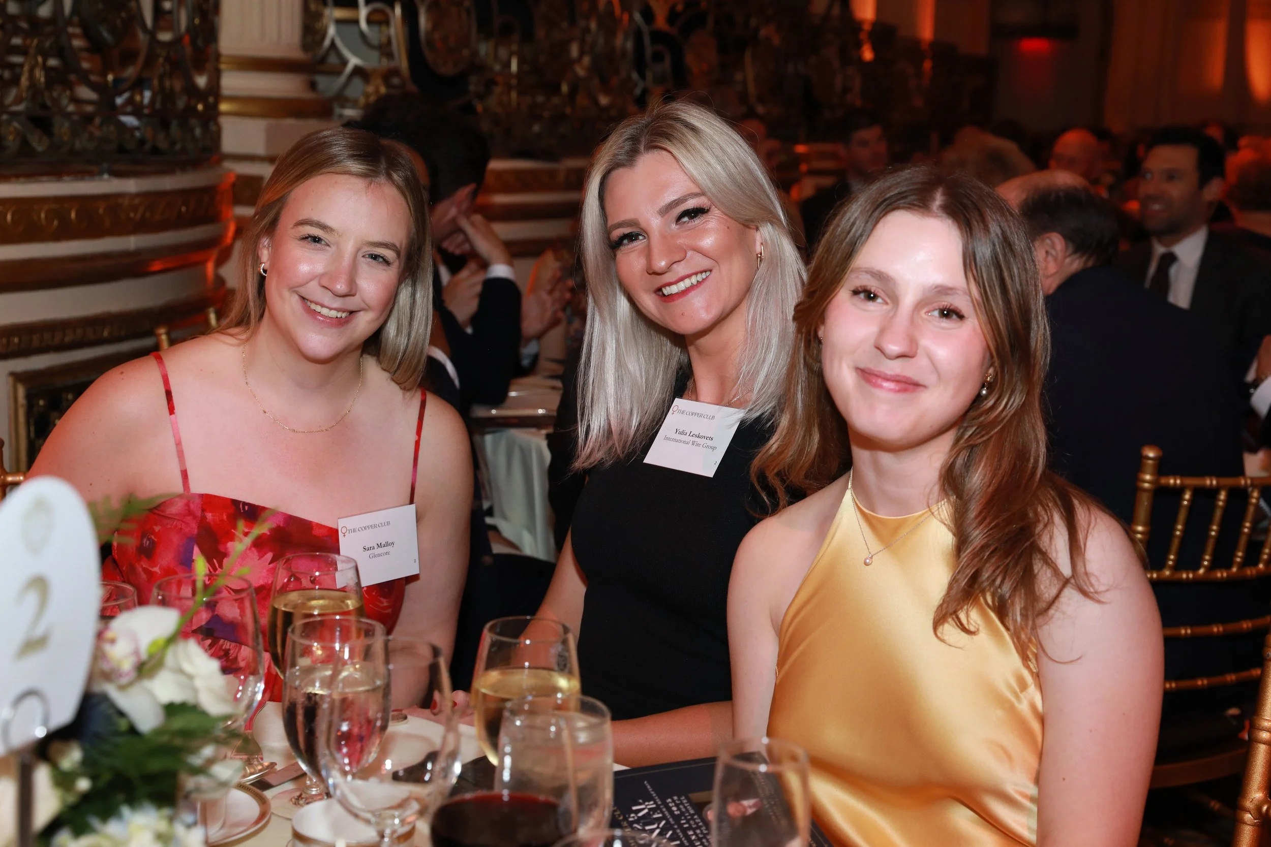 Three women celebrating at a formal event, smiling and sitting at a table with wine glasses, flowers, and menus in a decorated hall.