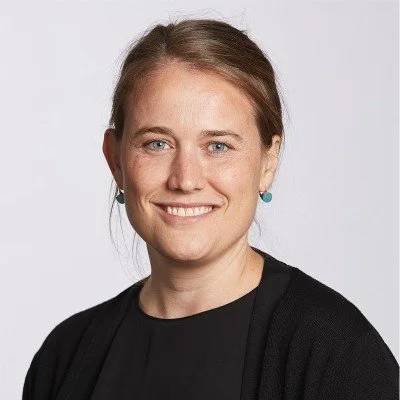 A woman with light brown hair in a ponytail, wearing a black top and turquoise earrings, smiling against a plain white background.