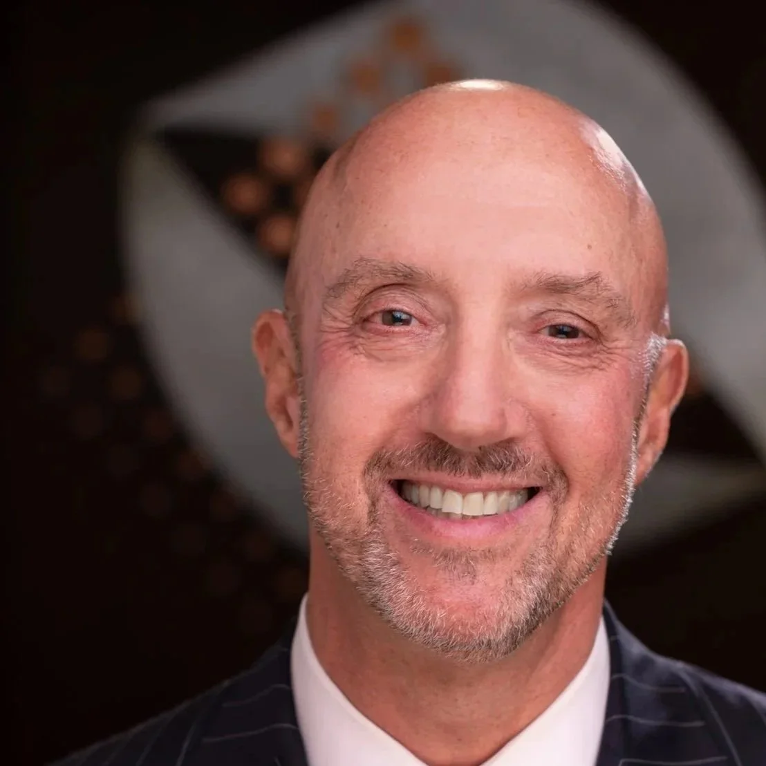 A smiling middle-aged man with a bald head and gray beard, wearing a pinstripe suit, in front of a blurred background.