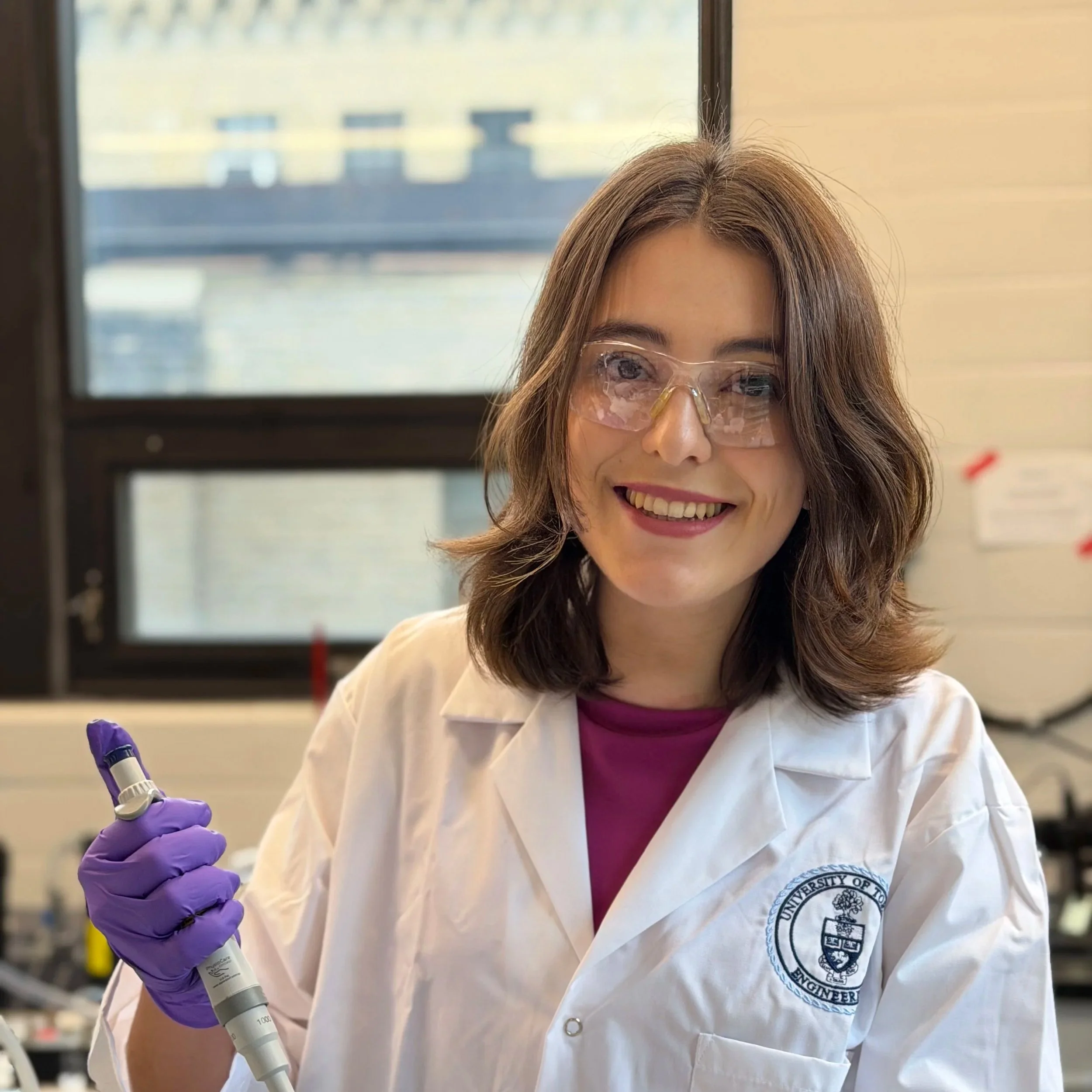 A woman with shoulder-length brown hair wearing glasses, a white lab coat with a university emblem, purple gloves, and holding a pipette in a laboratory setting.