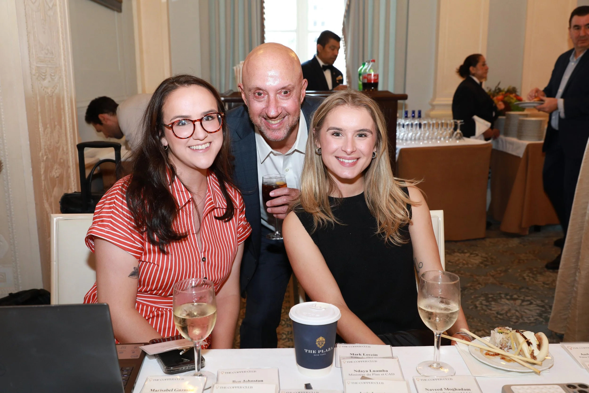 Three smiling people, two women and one man, posing at a formal event; they are seated at a table with name tags, drinks, and food, with others in the background in a decorated room.