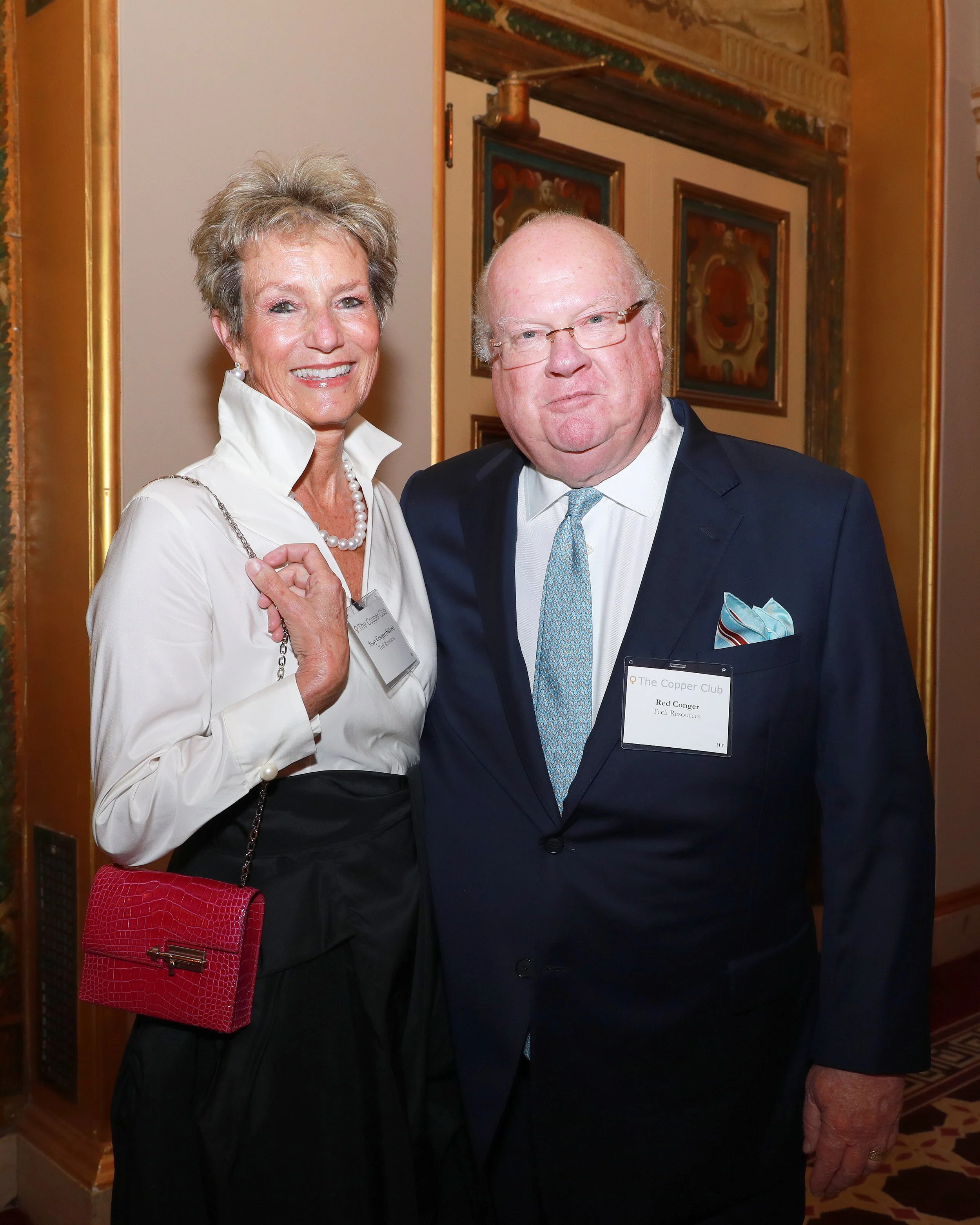 Two older adults standing together at a formal event. The woman on the left has short, styled blonde hair and is wearing a white collared shirt, pearl earrings, and a pearl necklace. She is holding a red textured handbag. The man on the right has bal