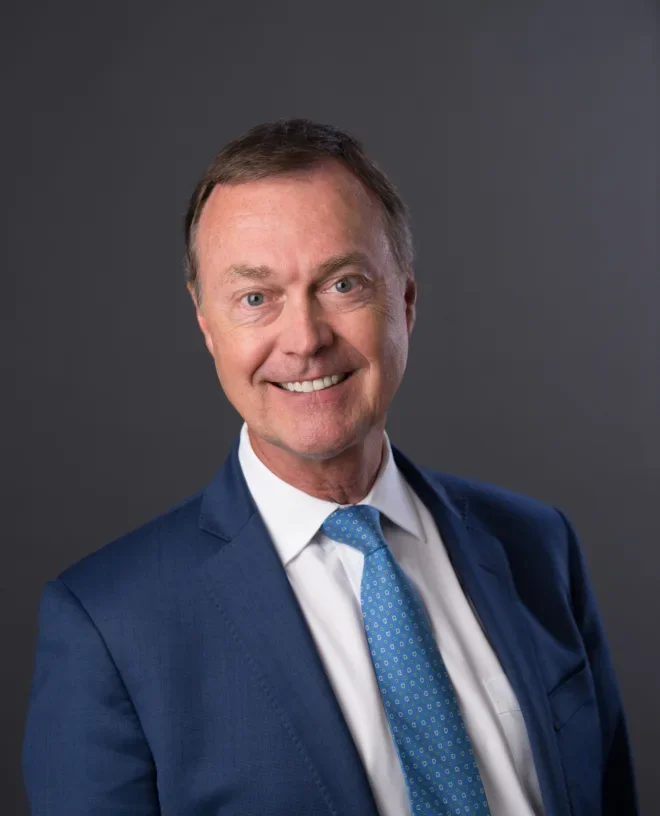 A middle-aged man with short brown hair smiling, dressed in a dark blue suit, white shirt, and a blue patterned tie, standing against a gray background.