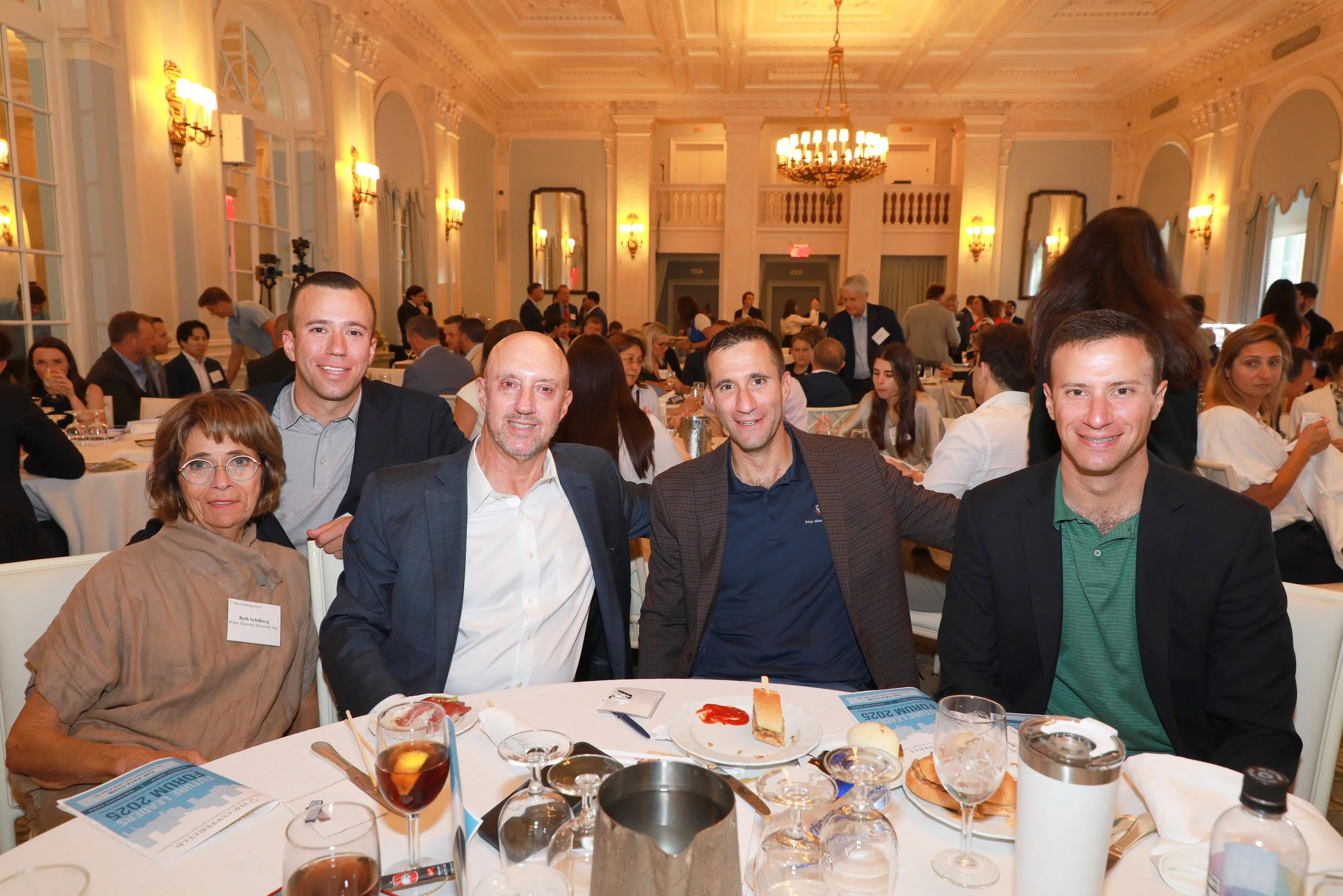 A group of five smiling people sitting at a round banquet table during a formal event in a large, elegant room with chandeliers and decorative lighting. Three men and two women are visible in the foreground, with food and drinks on the table.