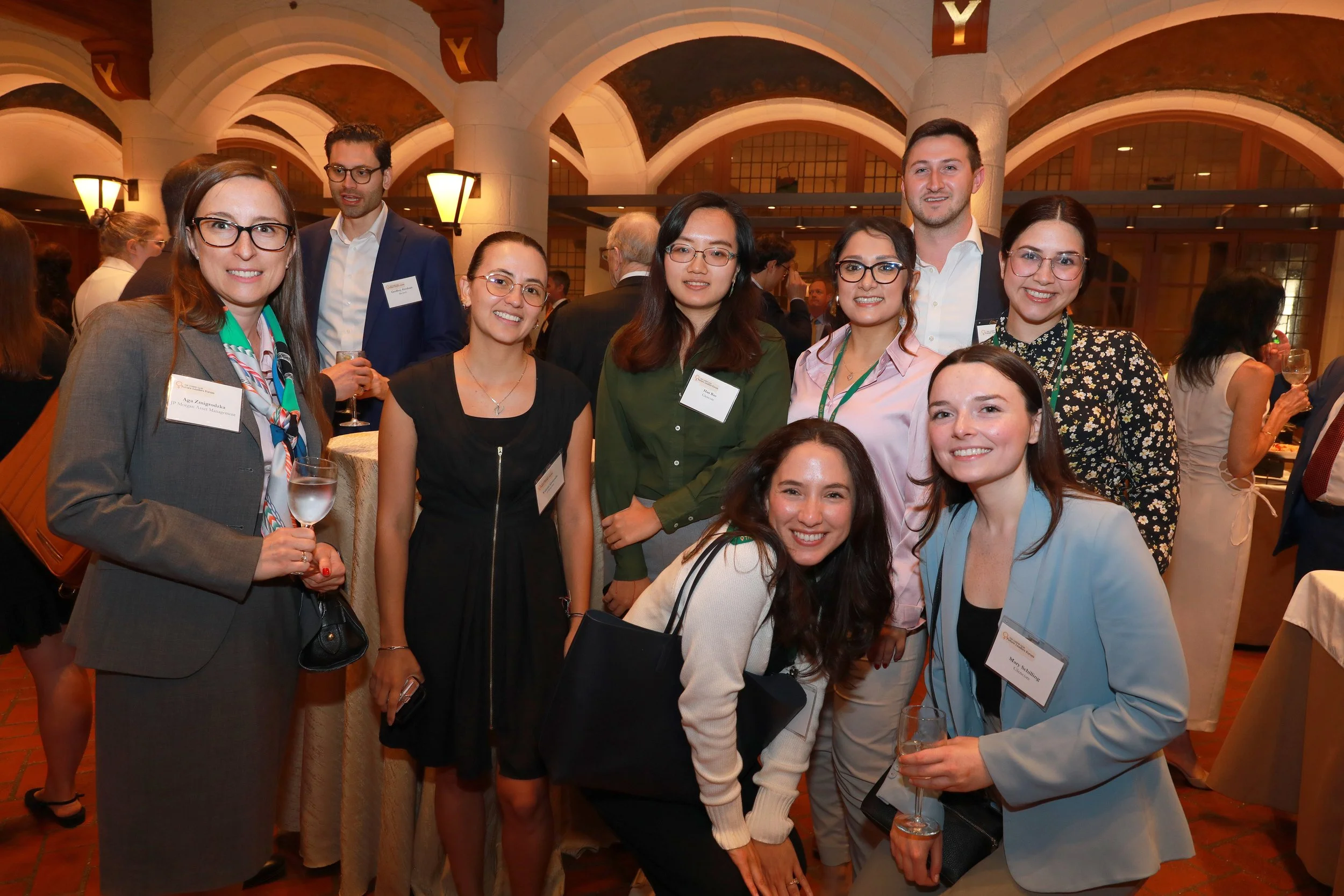 Group of professionally dressed people at an indoor networking event, smiling and holding drinks, in a warmly lit venue with high arched ceilings and decorative lighting.
