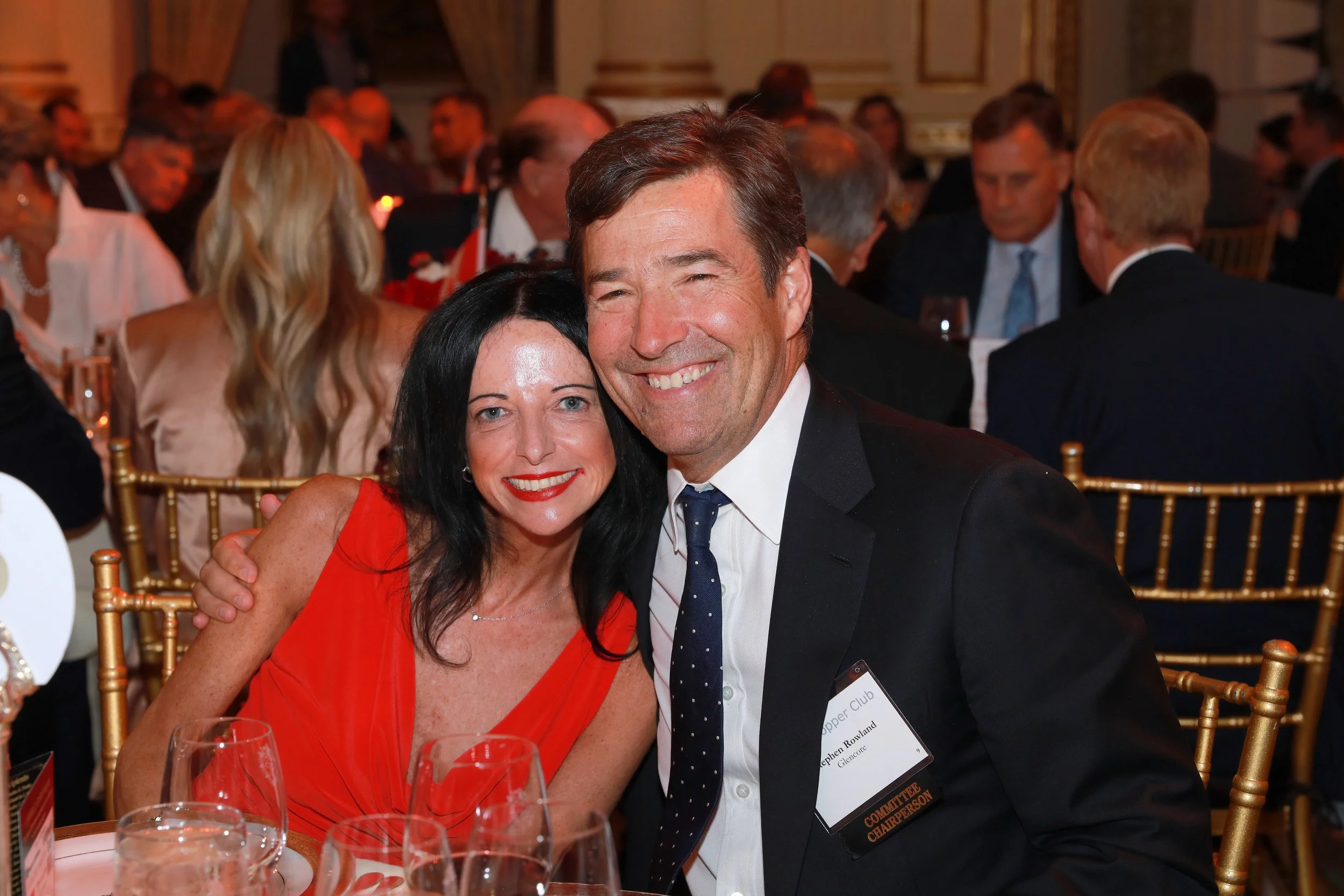 A man and woman are sitting together at a formal event, smiling at the camera. The woman has black hair and is wearing a red sleeveless dress. The man has brown hair, is wearing a dark suit with a white shirt and a dark tie, and has a name tag that r