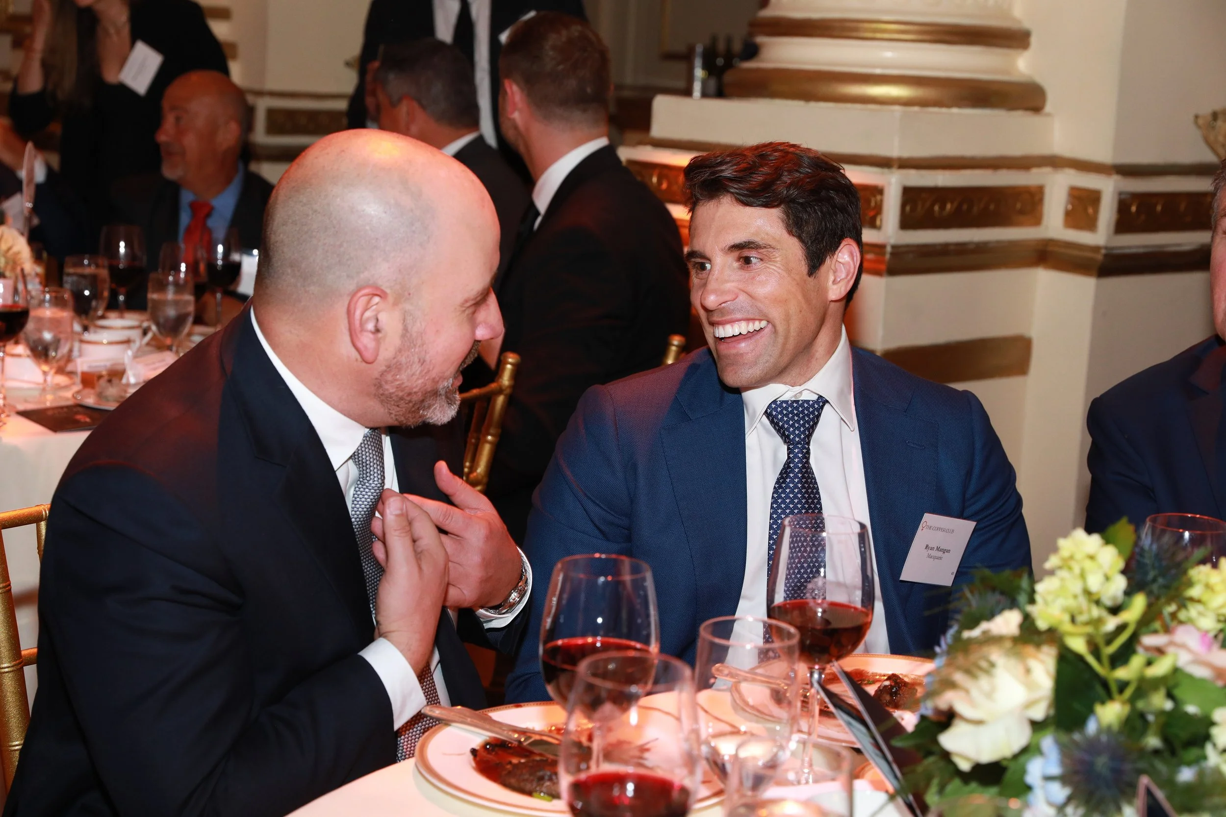 Two men dressed in suits are engaged in a conversation at a formal dinner event. The table is set with wine glasses, plates, and a floral centerpiece. Other guests can be seen in the background, and the decor features ornate, gold-accented architectu