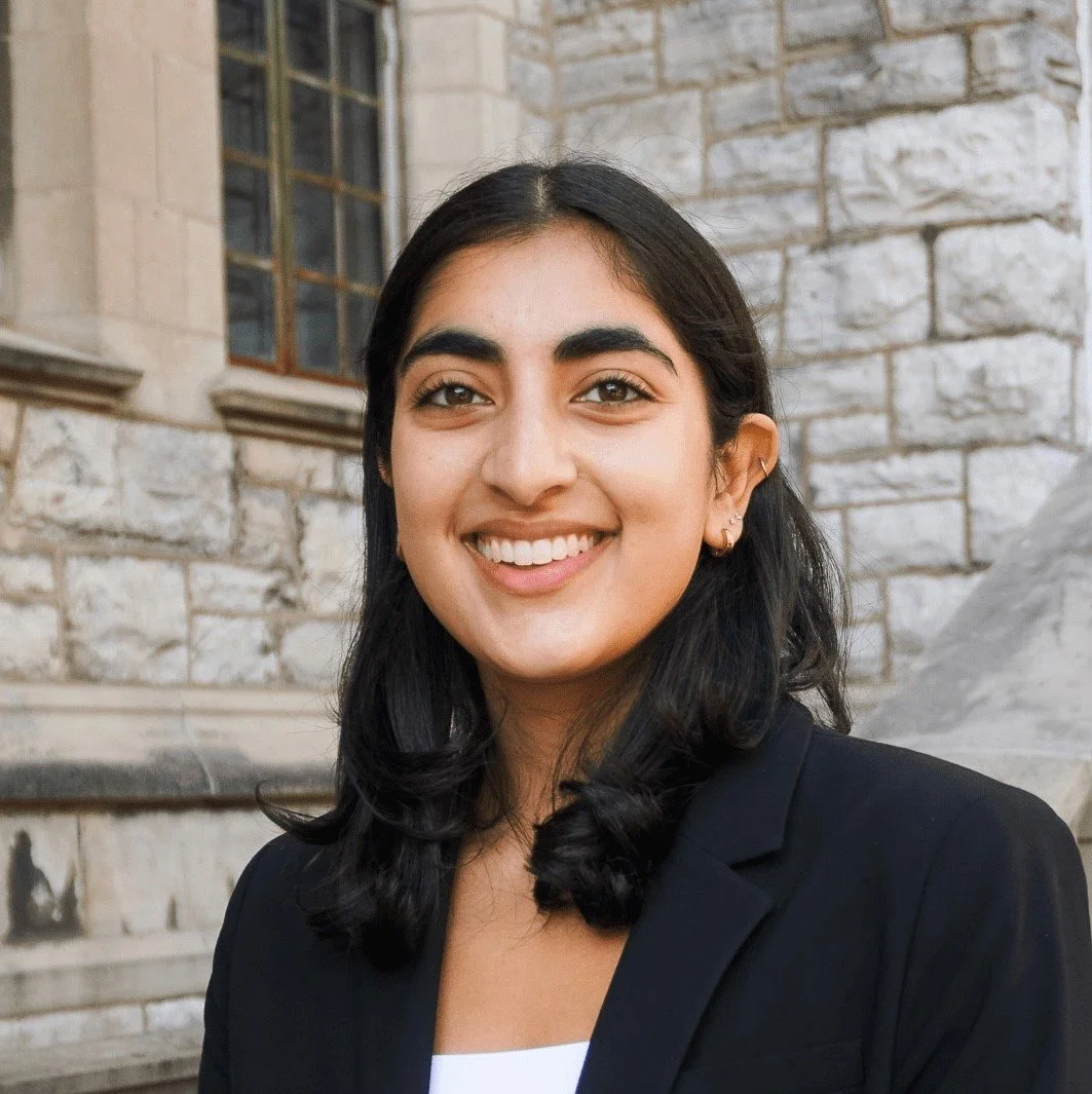 A young woman with dark hair and a bright smile, wearing a black blazer, standing in front of a stone building with large windows.