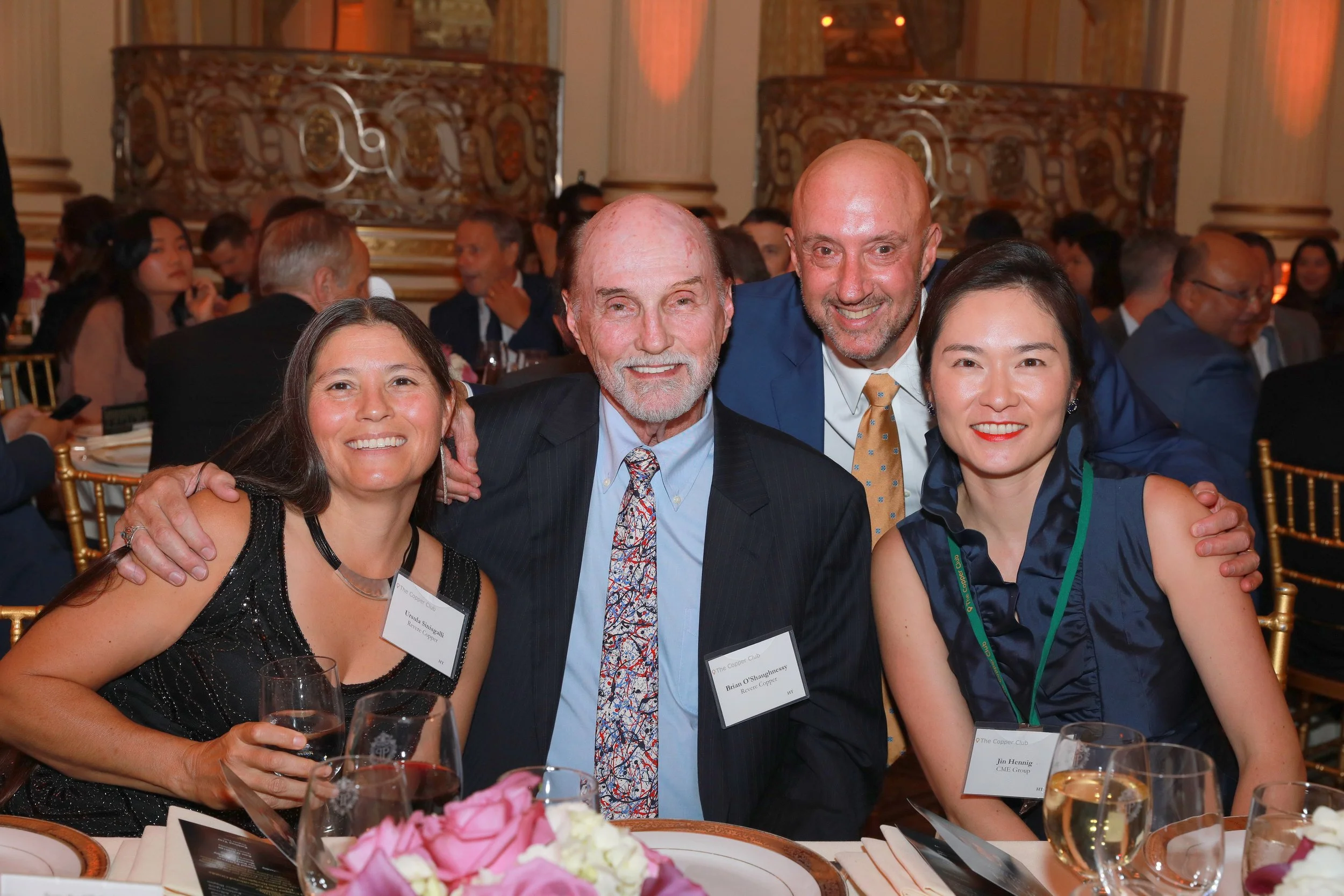 Four people sitting at a dinner table in formal attire at an event, smiling for the camera, with a decorative background and other guests seated in the background.