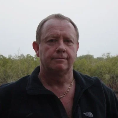 A middle-aged man with short, light brown hair and a slight smile, wearing a black jacket and a gold chain, stands outdoors with trees and overcast sky in the background.