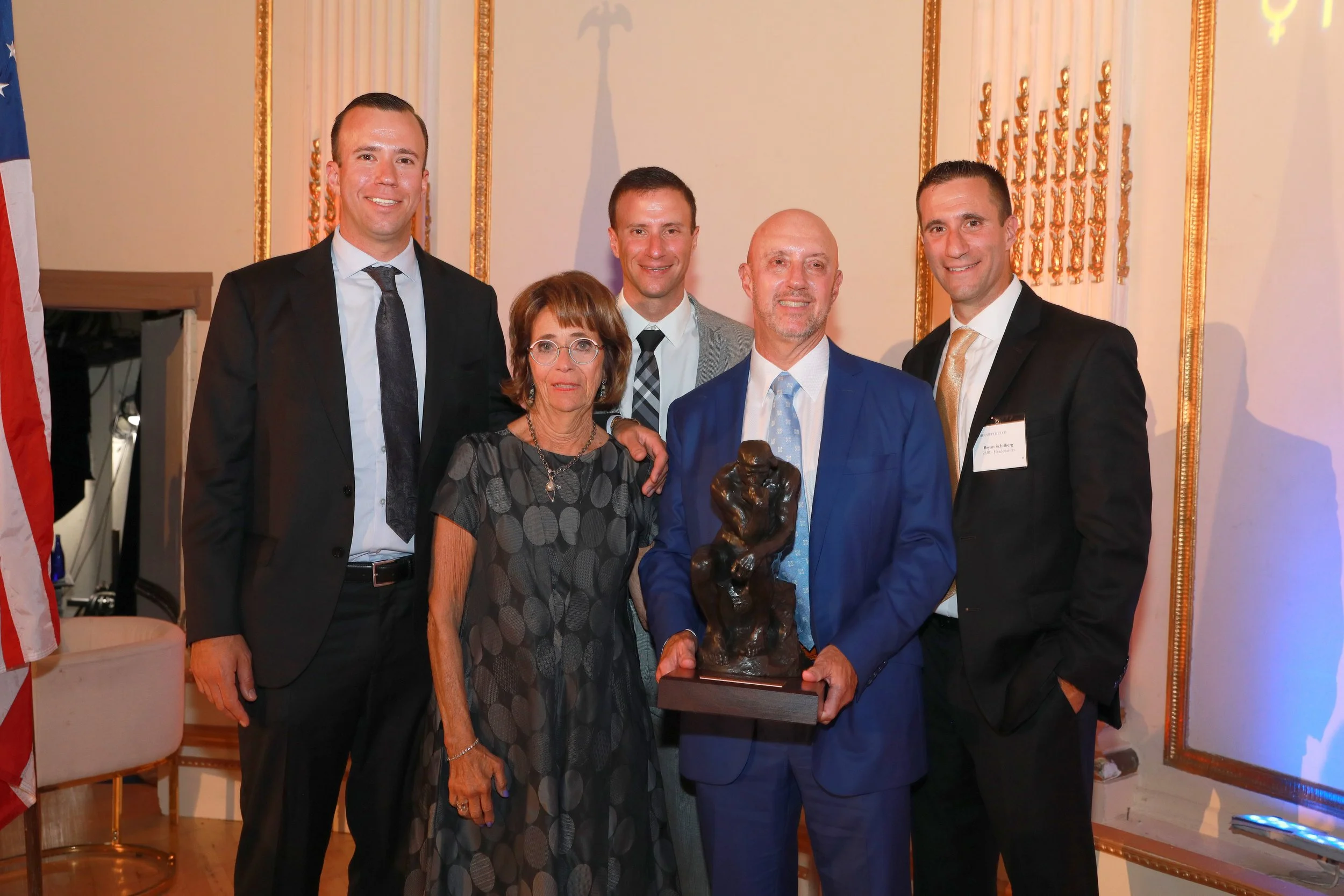 A group of six people, including four men in suits and two women, pose together in a decorated formal setting. One man is holding an abstract sculpture or statue. They are standing in front of a wall with ornate gold accents and a large abstract shad