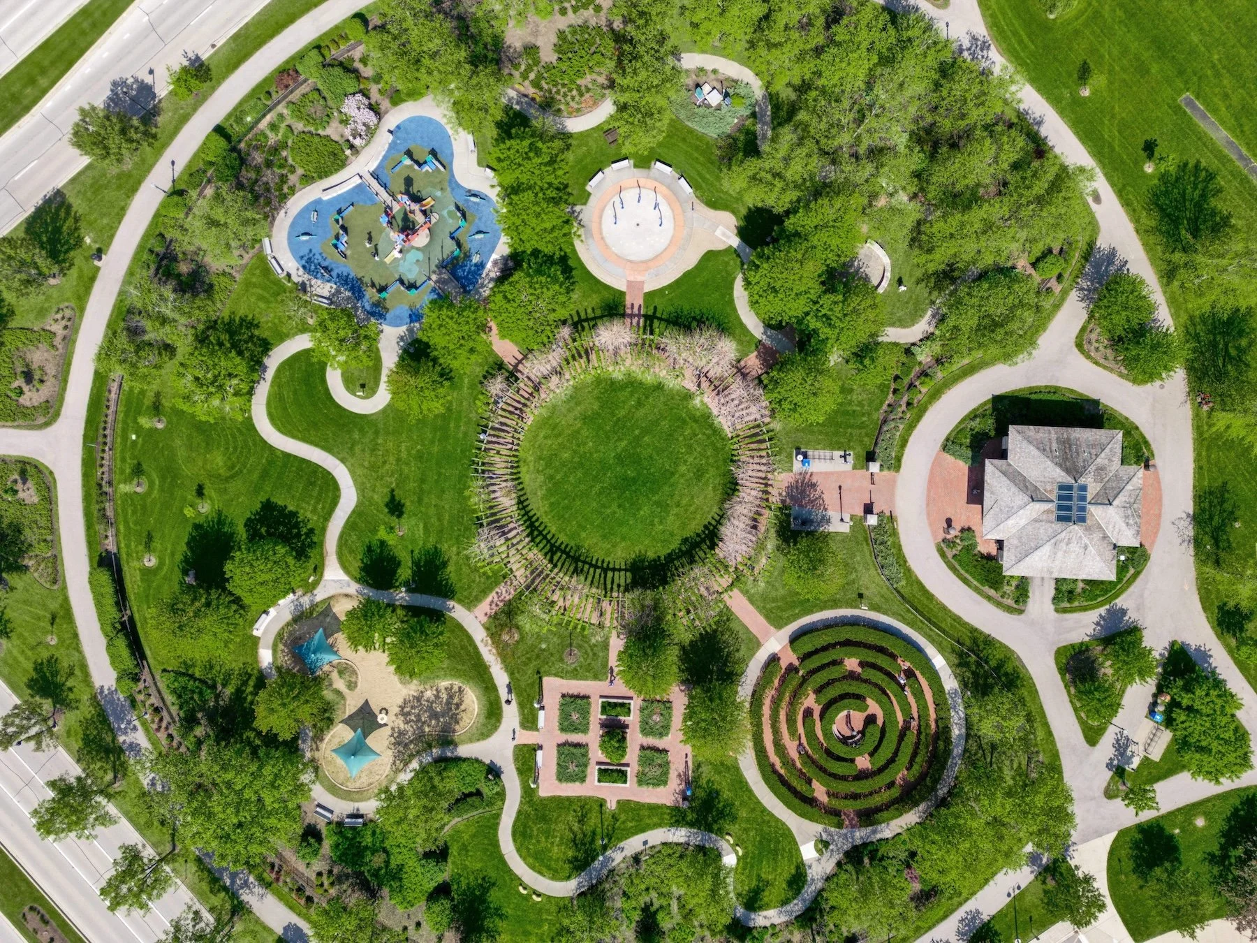 Aerial view of a park with winding pathways, green lawns, trees, playgrounds, a gazebo, and surrounding roads.