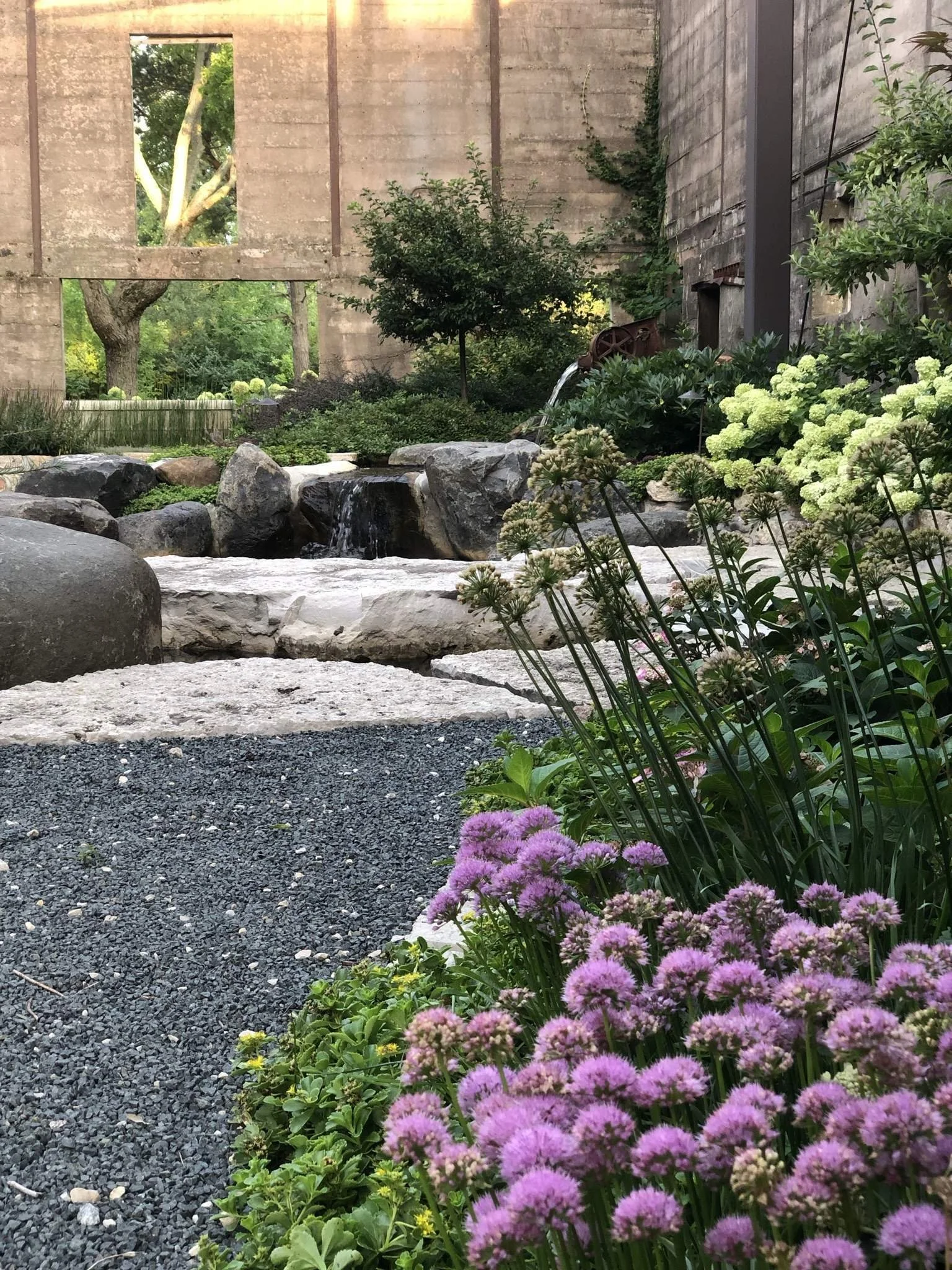 A garden with purple flowers in the foreground, a small waterfall with rocks, green shrubs, and trees, and an unfinished concrete building in the background.
