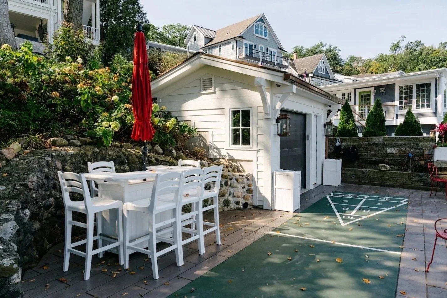 Backyard patio with white high-top table and chairs, red umbrella, a shuffleboard court, and a small white shed surrounded by houses and greenery.