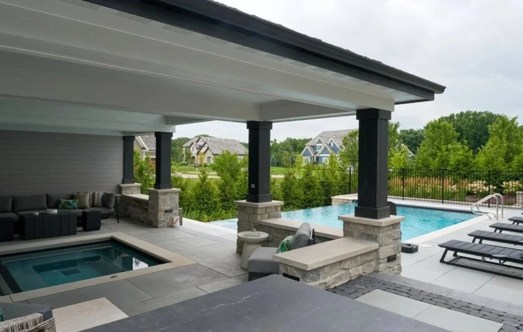 Outdoor patio with a covered seating area, a hot tub, a swimming pool, and lounge chairs, surrounded by trees and neighboring houses