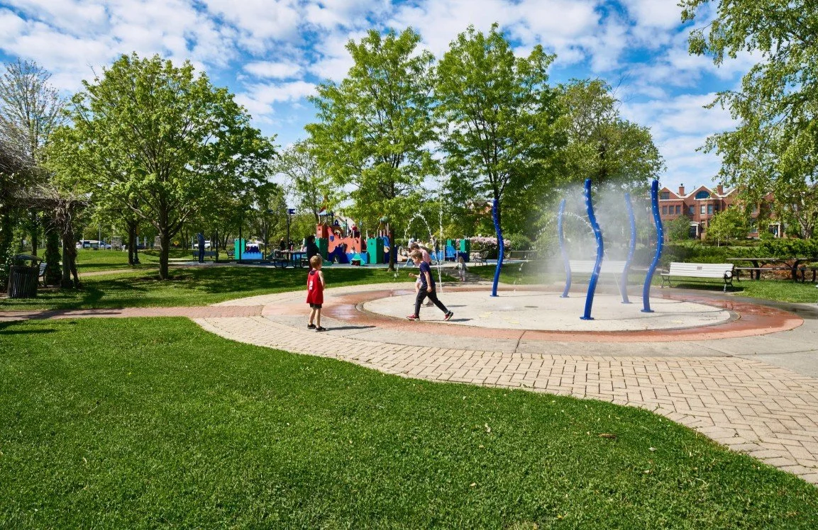 Children playing around water spray fountain in a park with trees, playground, benches, and houses in the background on a sunny day.